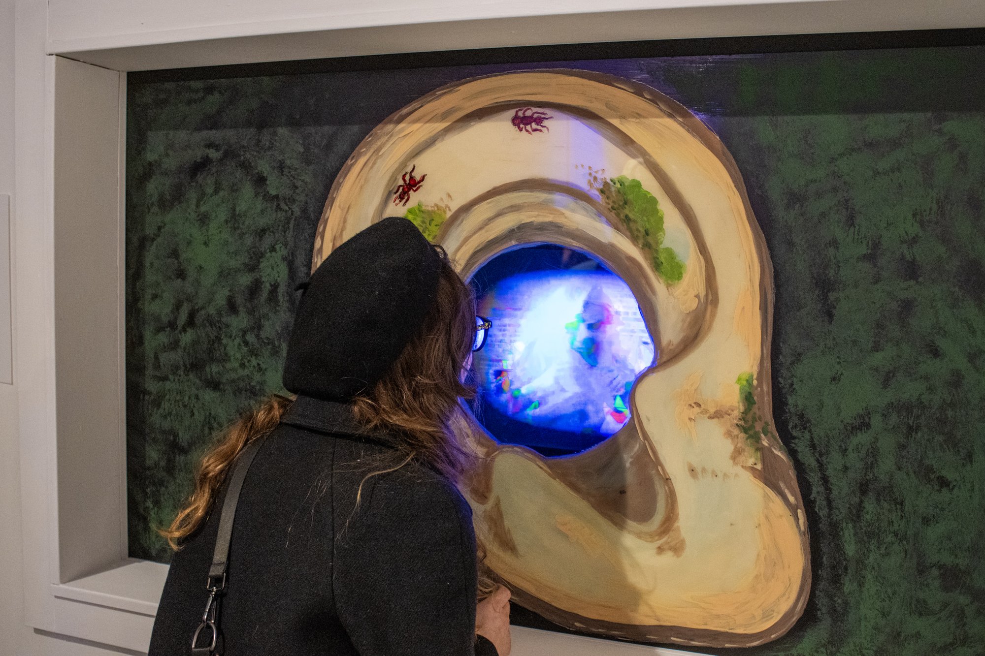 Woman looking through a window with painted ear and painted grass that creates a window where she is looking at a TV screen in Uncanny Art House