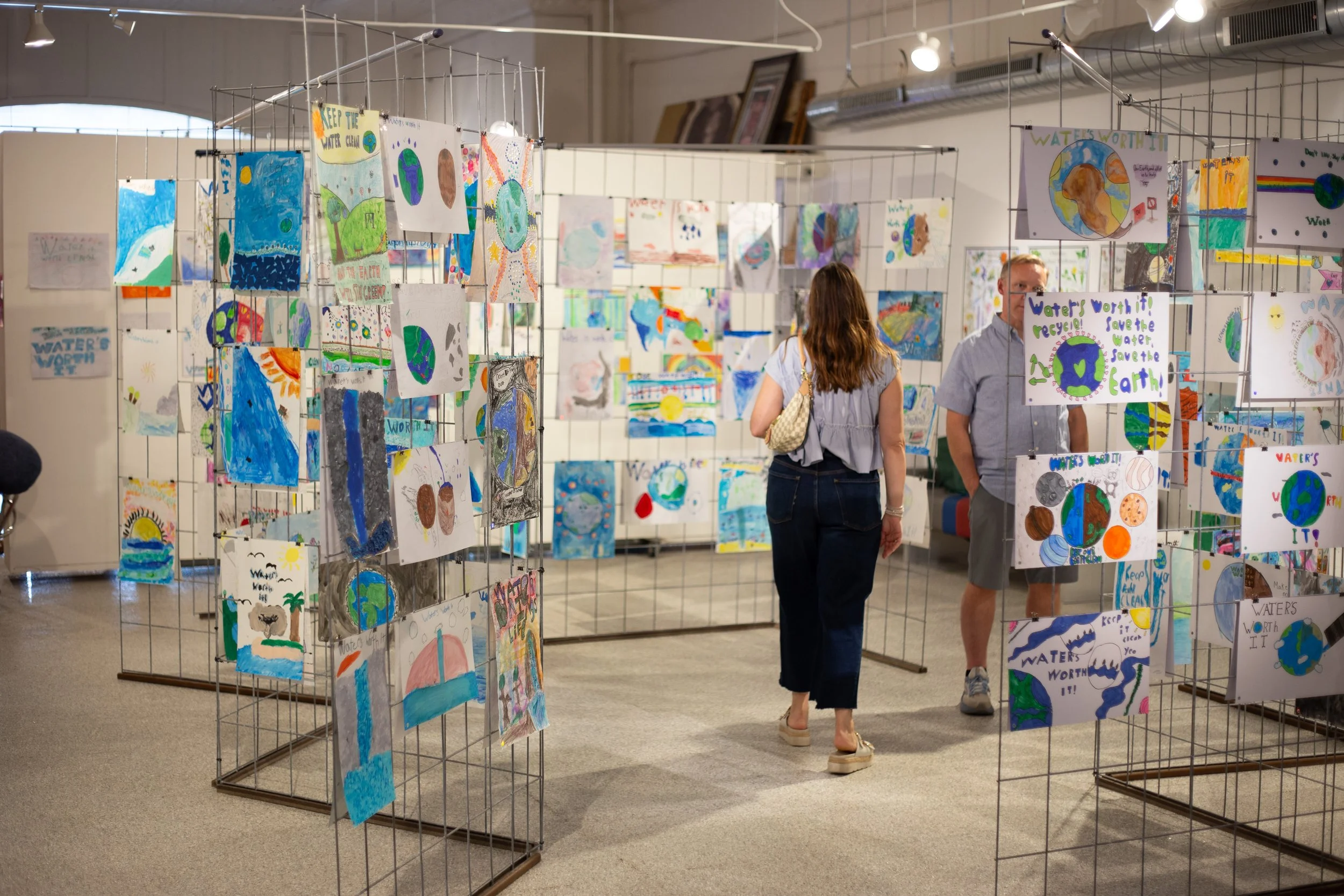 Two people walking in an art gallery surrounded by art submissions from elementary students