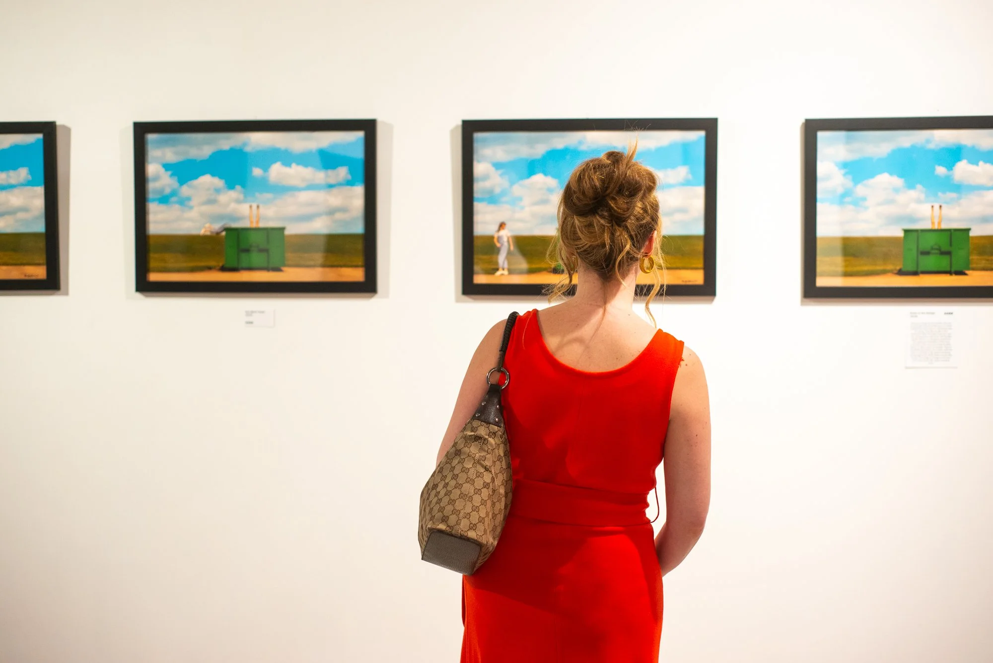 Gallery guest in a red dress looks at a series of brightly colored photographs on the wall including a sequence of blue skies, white clouds, and a green dumpster with a woman upside down and legs positioned like a mannequin and another woman walking