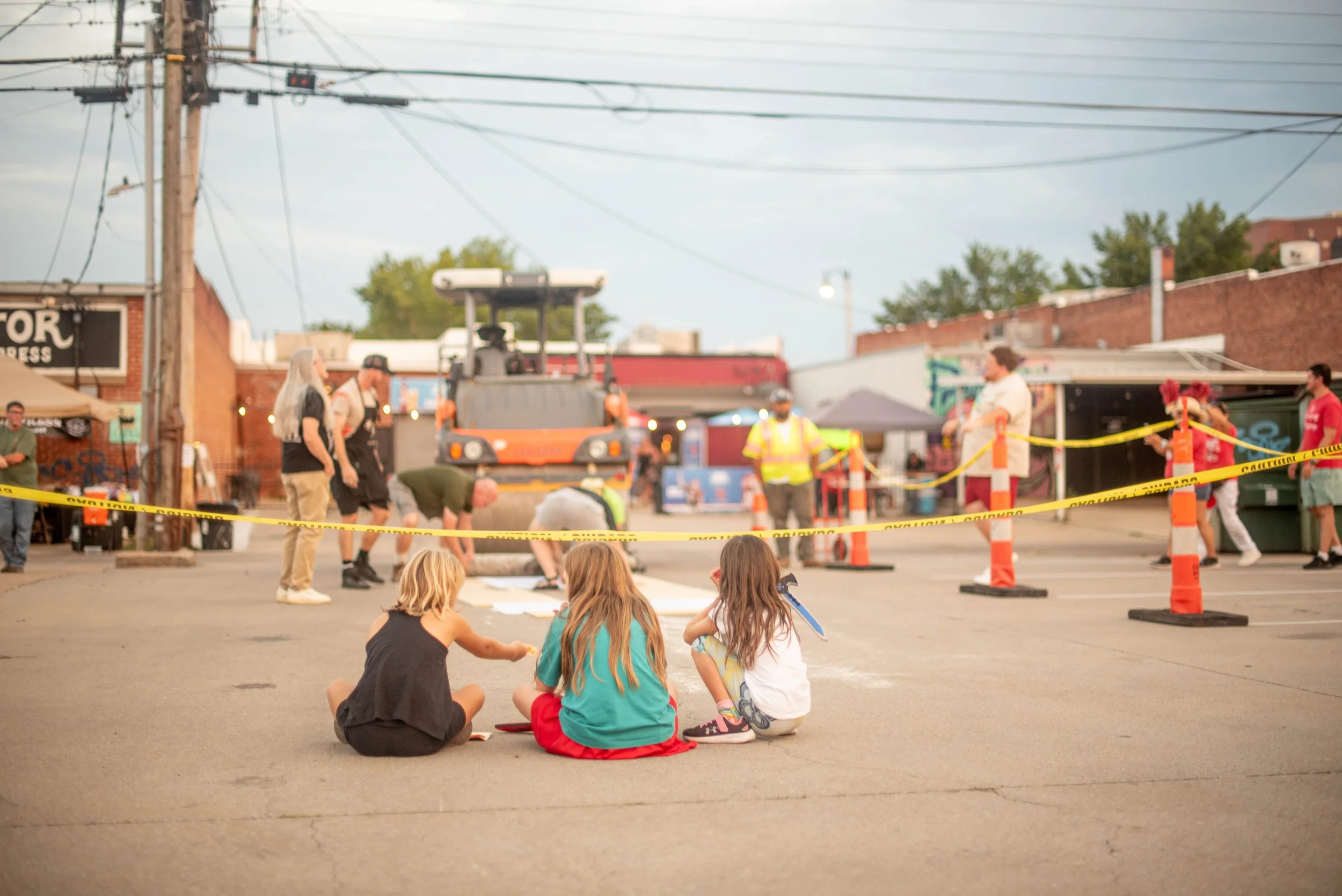 Three children sitting infront of the steamroller area watching prints be pulled to hang with several people watching