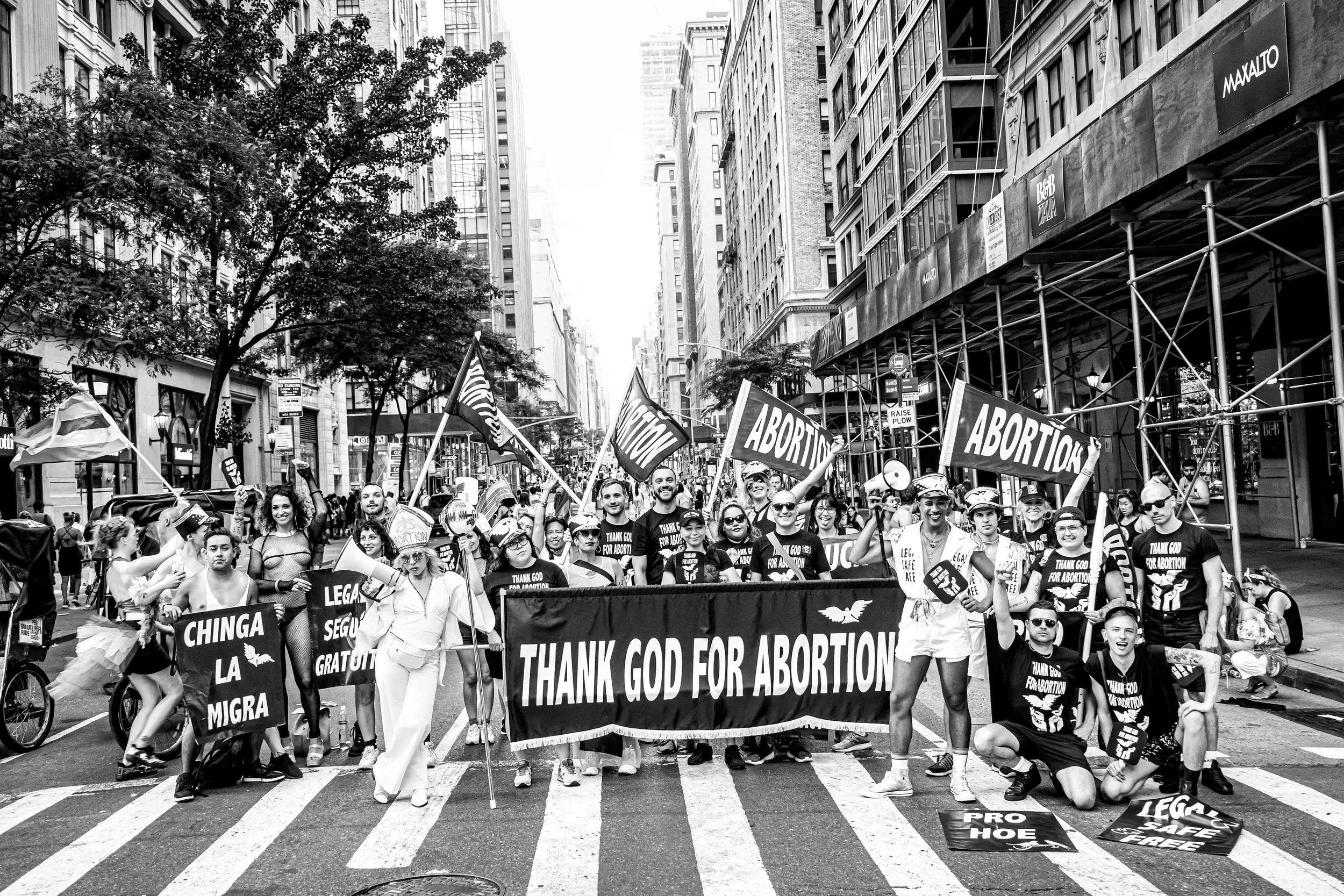 A black-and-white photograph shows a crowd at the 2015 NYC Pride Parade. People carry signs including “Chinga La Migra,” “Abortion,” and “Pro Hoe,” while two people hold a “THANK GOD FOR ABORTION” banner.