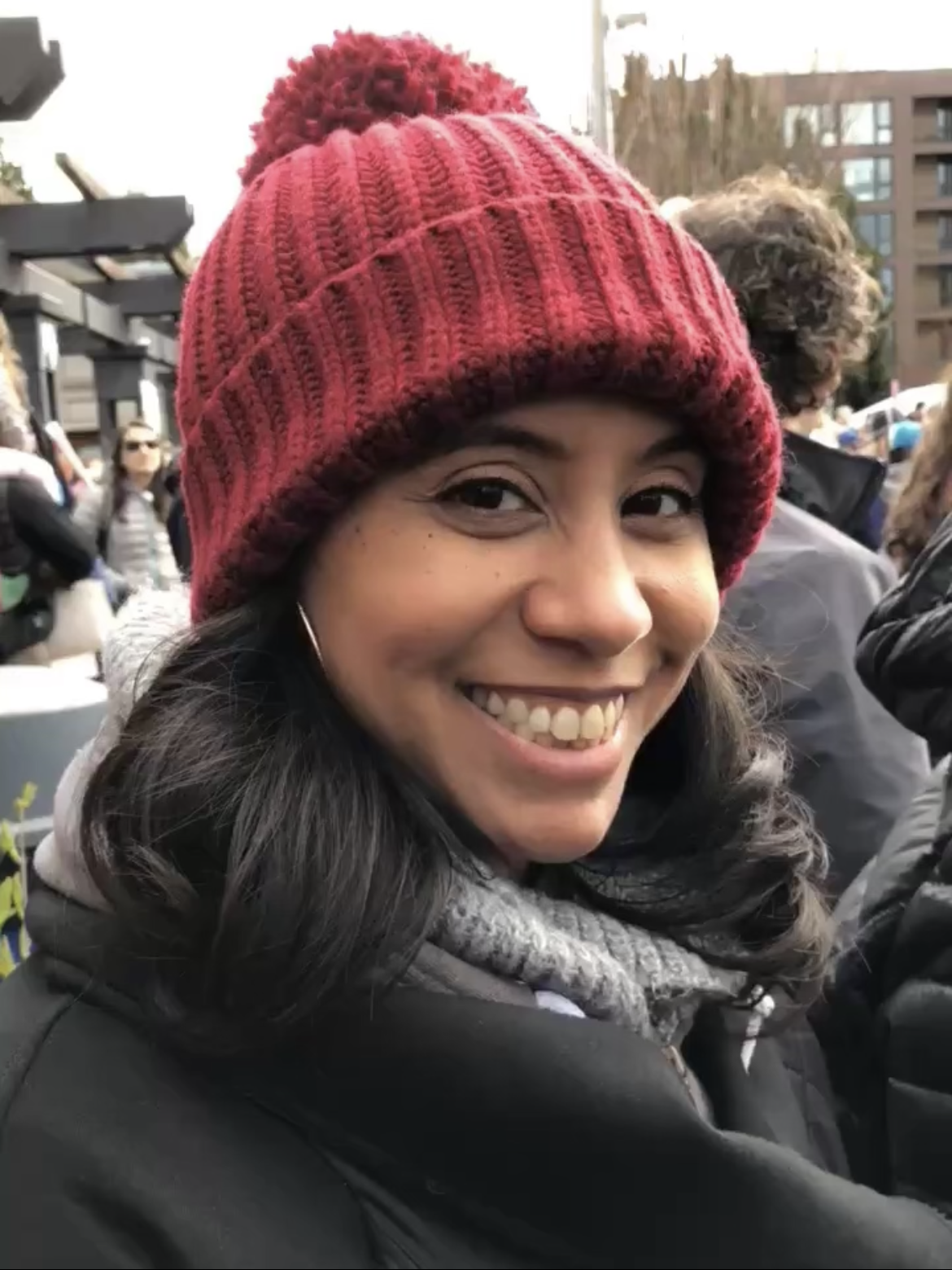 Smiling woman wearing a red knit hat with a pom-pom, black jacket, and gray scarf, outdoors at a crowded event.