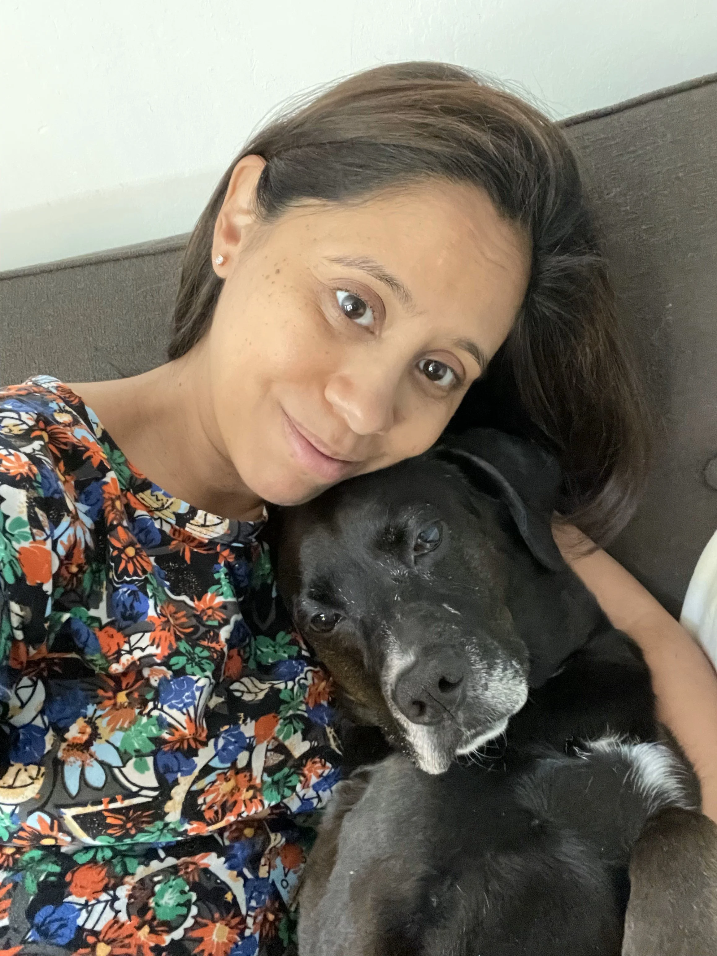 A woman with light brown hair and small earrings lying on a couch, snuggling with a black dog that has a white patch on its nose. The woman is wearing a floral shirt and smiling softly.