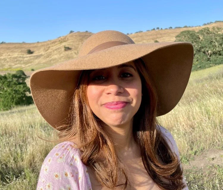 Young woman with long hair wearing a large tan hat, standing outdoors in a grassy field with hills behind her on a sunny day.