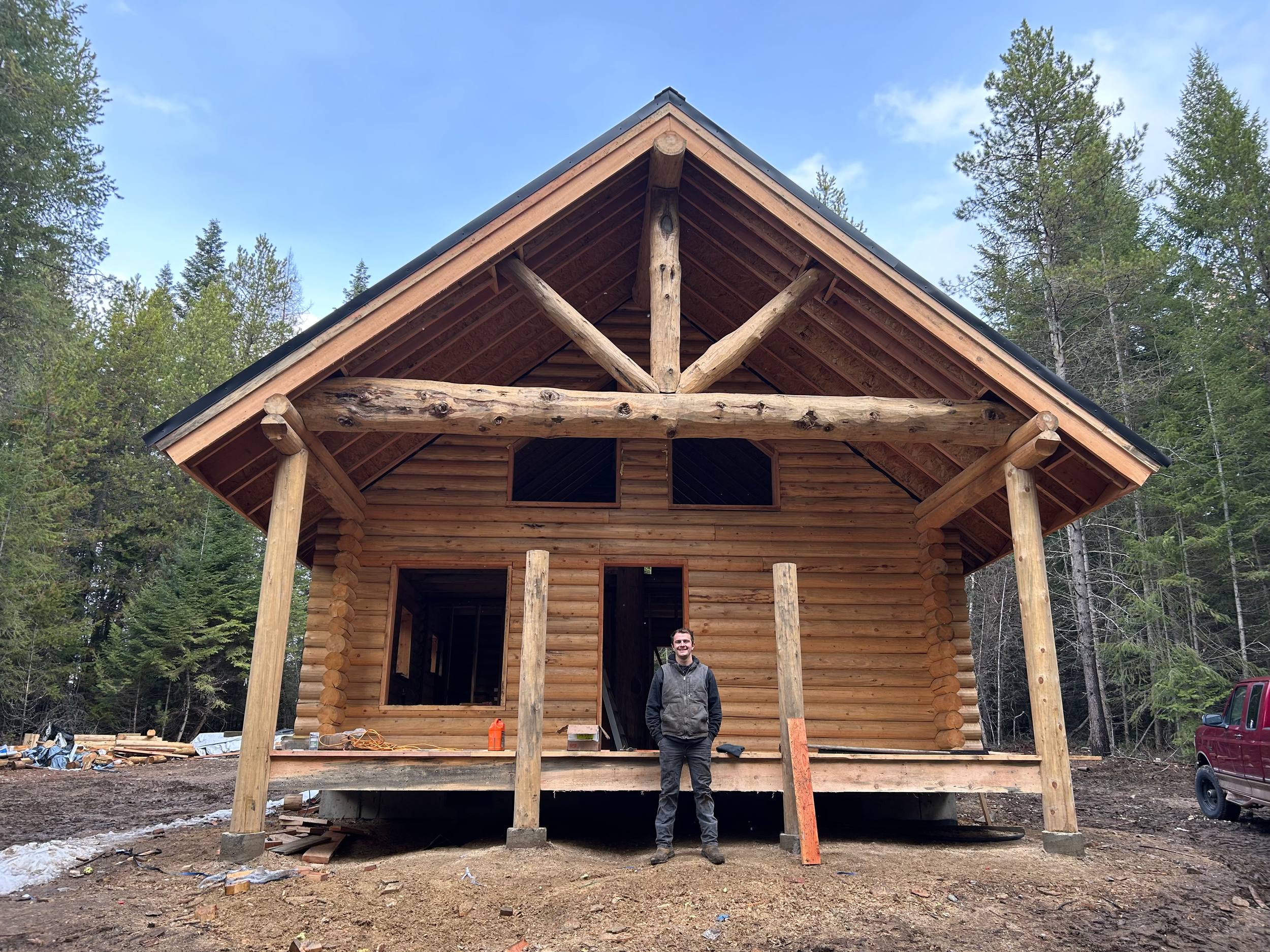 A man stands in front of a partially constructed wooden house in a forested area, with tall trees surrounding the site and construction materials scattered on the ground.
