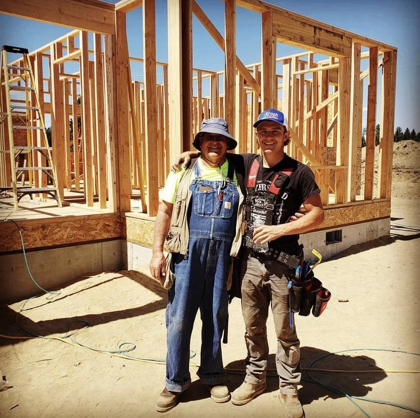 Two construction workers, one older and one younger, standing in front of a wooden house frame under construction on a sunny day. They are smiling, with the older man wearing overalls and a bucket hat, and the younger man wearing a cap and carrying tool pouches.