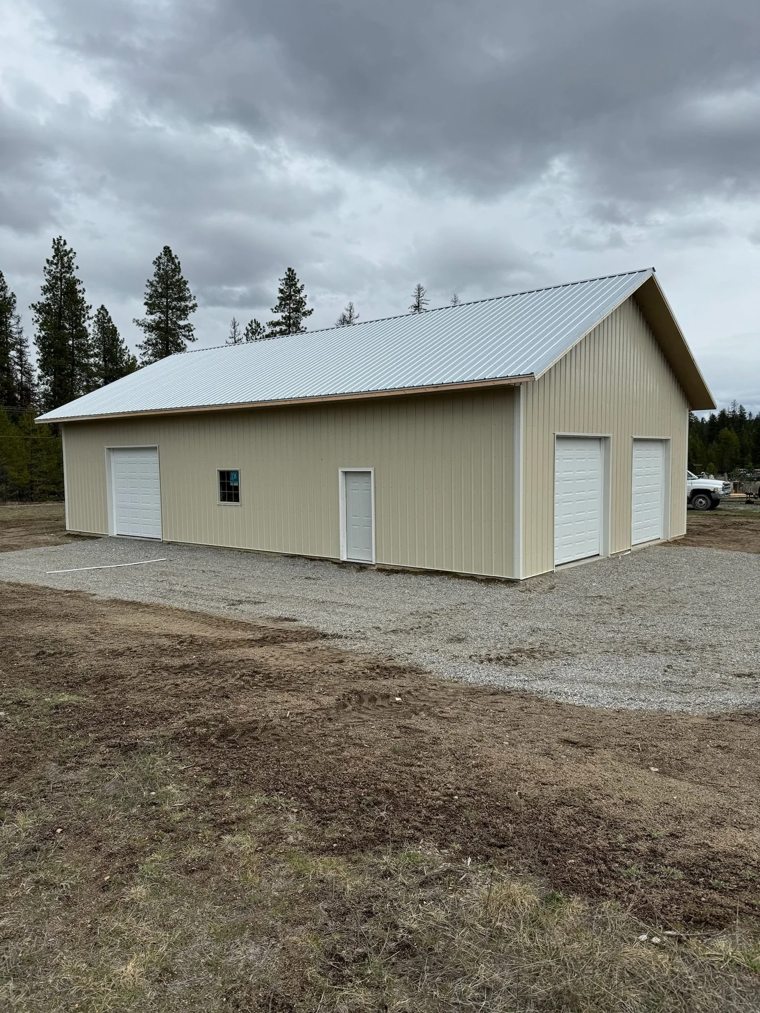 A beige metal building with three garage doors and one door on the side, situated on a dirt lot with grass in the foreground. A cloudy sky and tall pine trees are in the background.