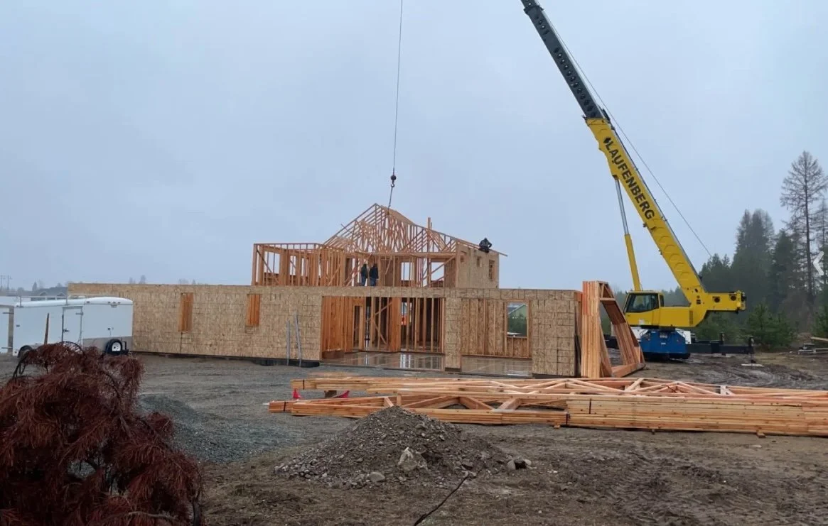 A house under construction with wooden framing, a crane lifting a part of the structure, construction materials on the ground, and an overcast sky.