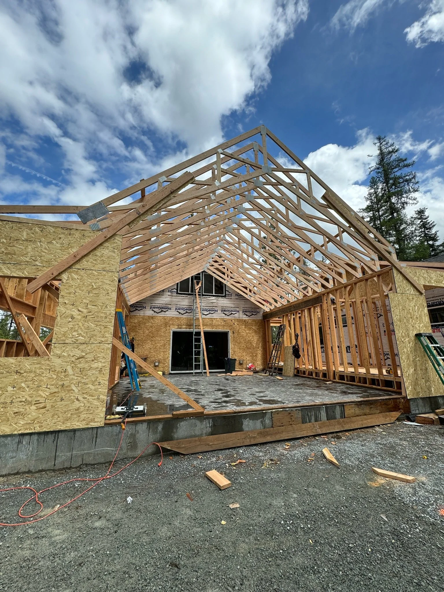 Construction site where a house is being built, showing the wooden frame and roof structure in progress against a cloudy sky.