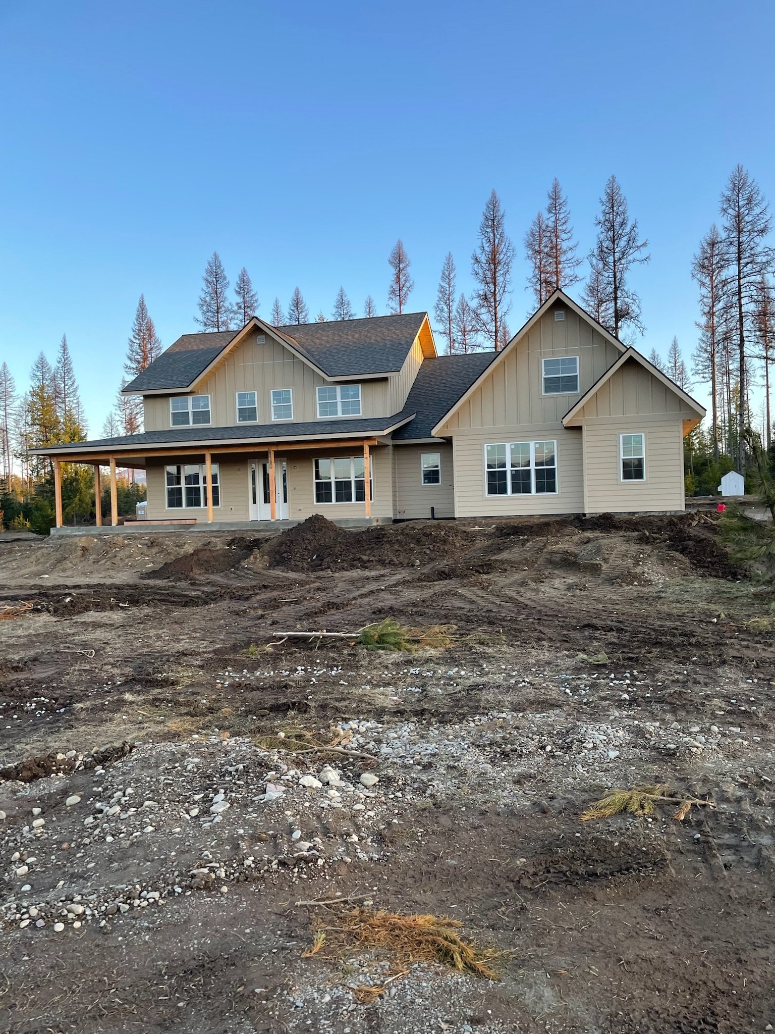 Newly constructed house with beige siding, black roof, and white window frames, set on a dirt yard with some scattered rocks and a small pile of dirt in the foreground. Tall trees are in the background under a clear blue sky.