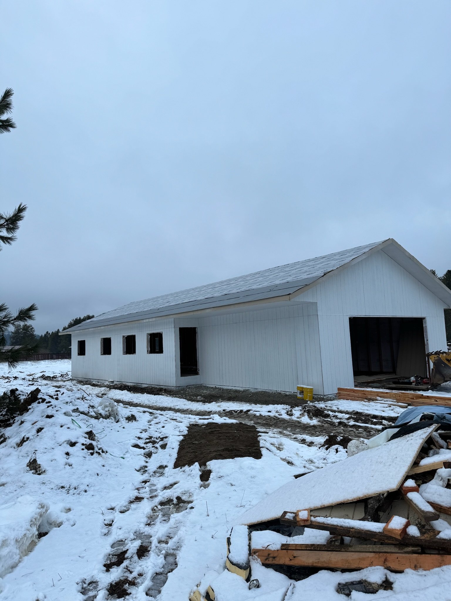 Construction site with a white building under construction during winter, snow on the ground, construction materials scattered, and trees in the background.