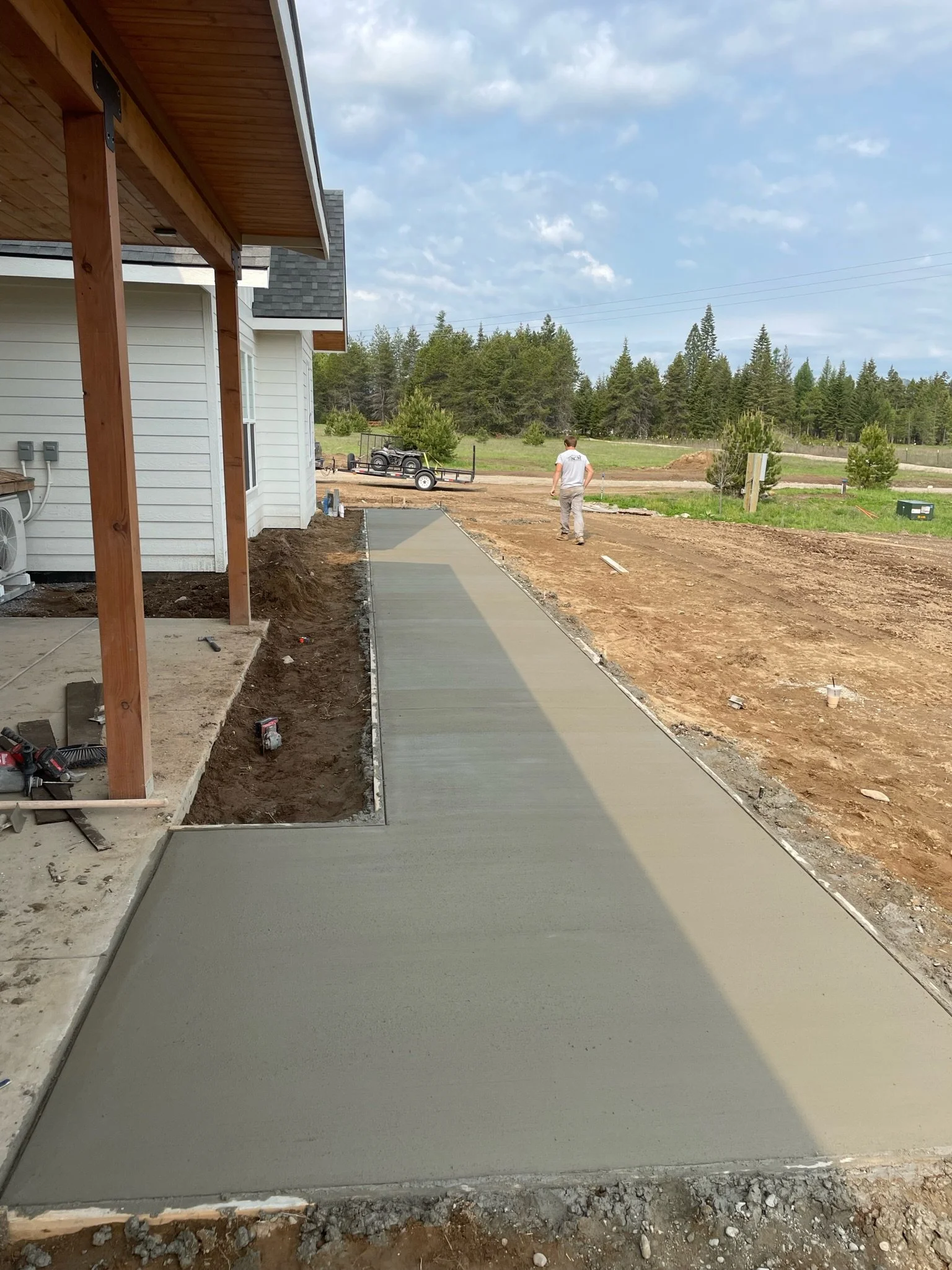 A newly poured concrete sidewalk next to a house under construction, with a man walking on the dirt beside it. In the background, there is a trailer and trees under a partly cloudy sky.