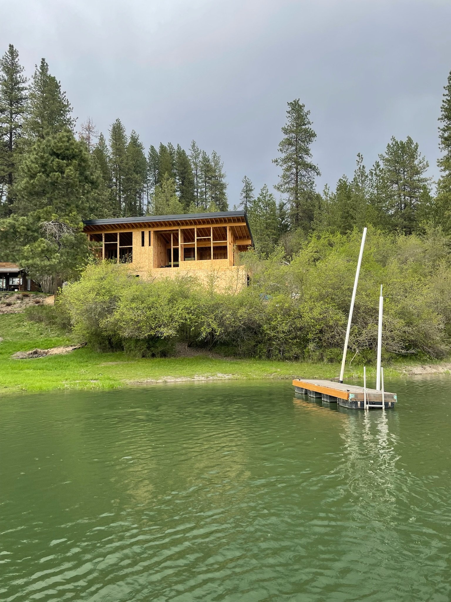 Construction of a house with a wooden frame on a hillside overlooking a body of water, with a small dock and green trees.