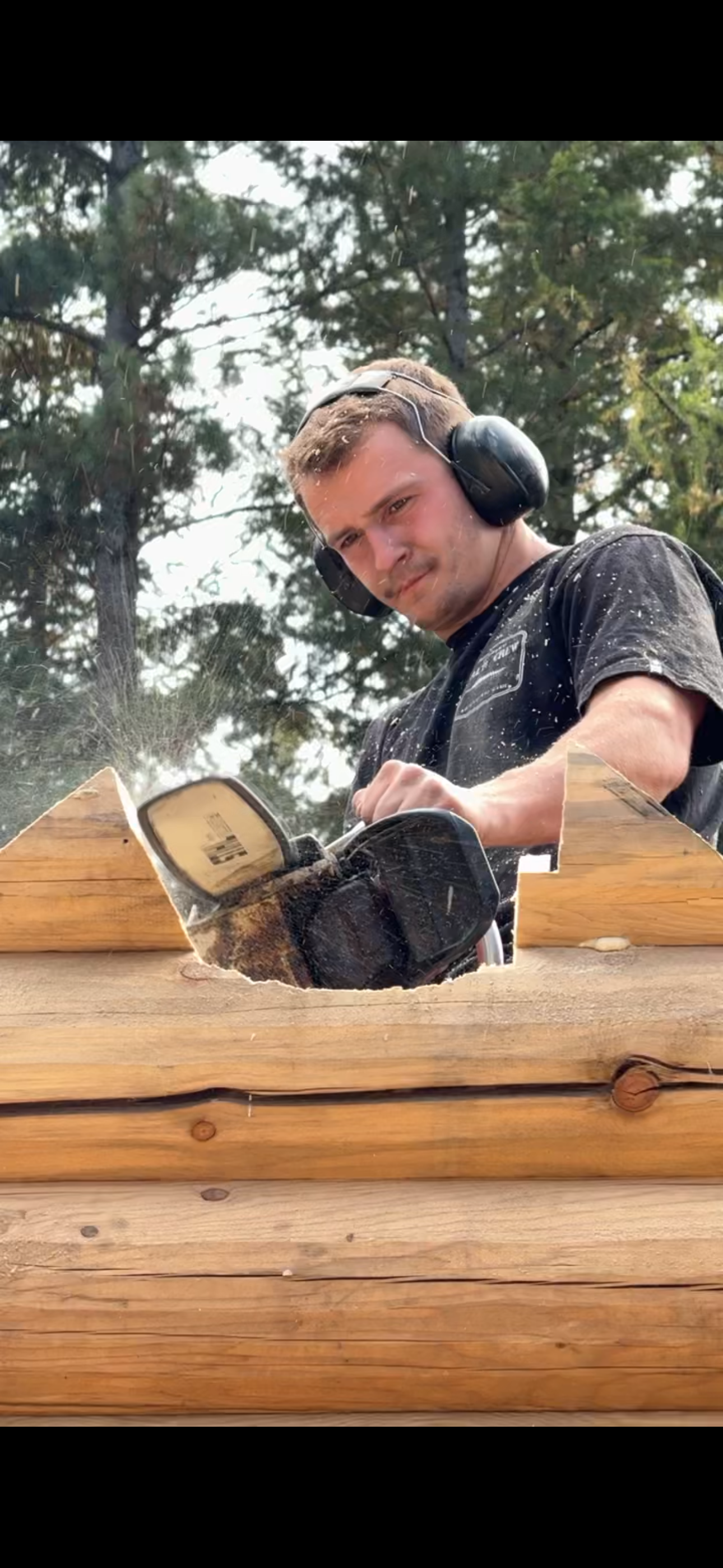 Man cutting wood with a power saw outdoors, wearing hearing protection, with trees in the background.