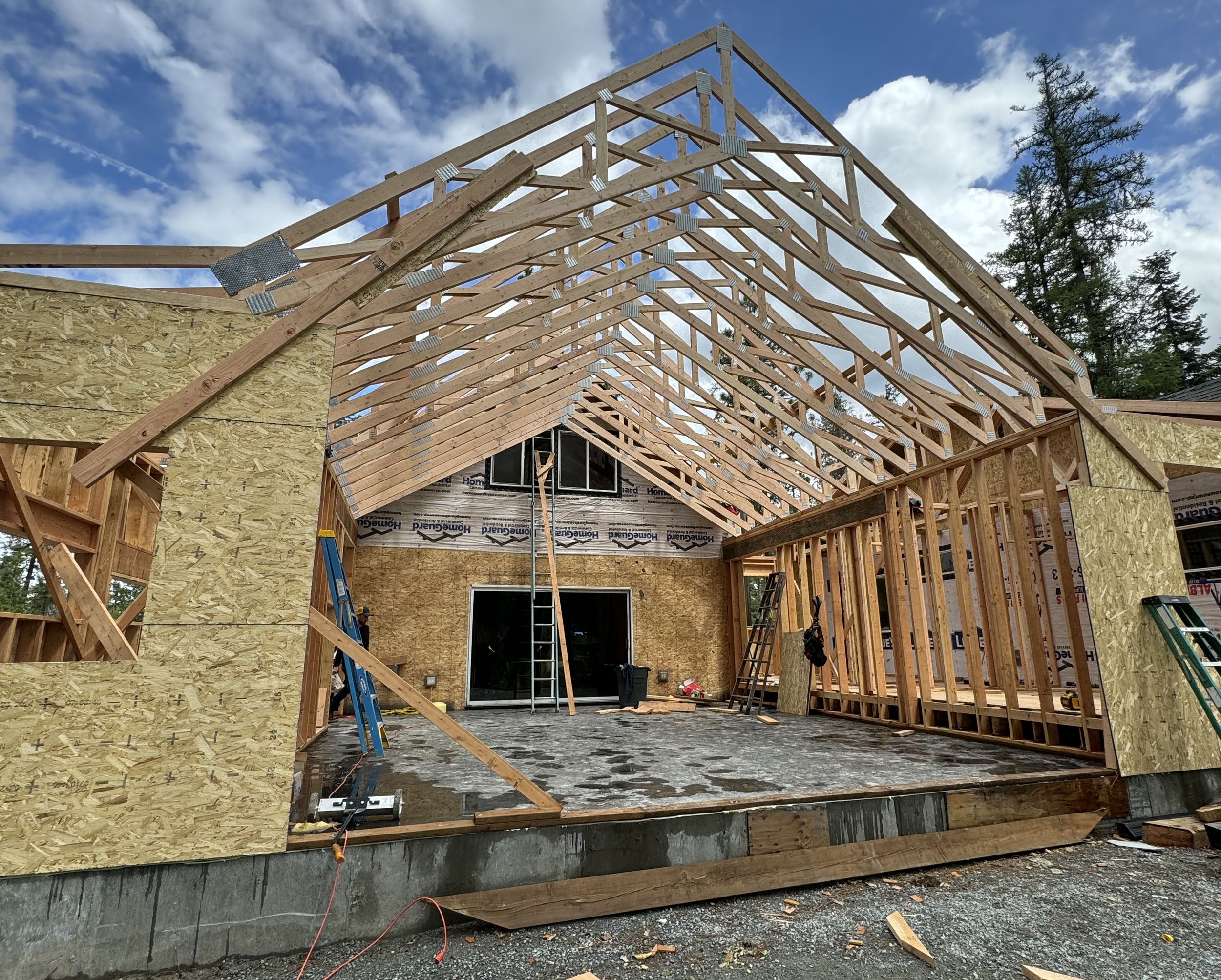 Construction site with a house frame, wooden roof trusses, and plywood walls against a bright sky with clouds and trees.