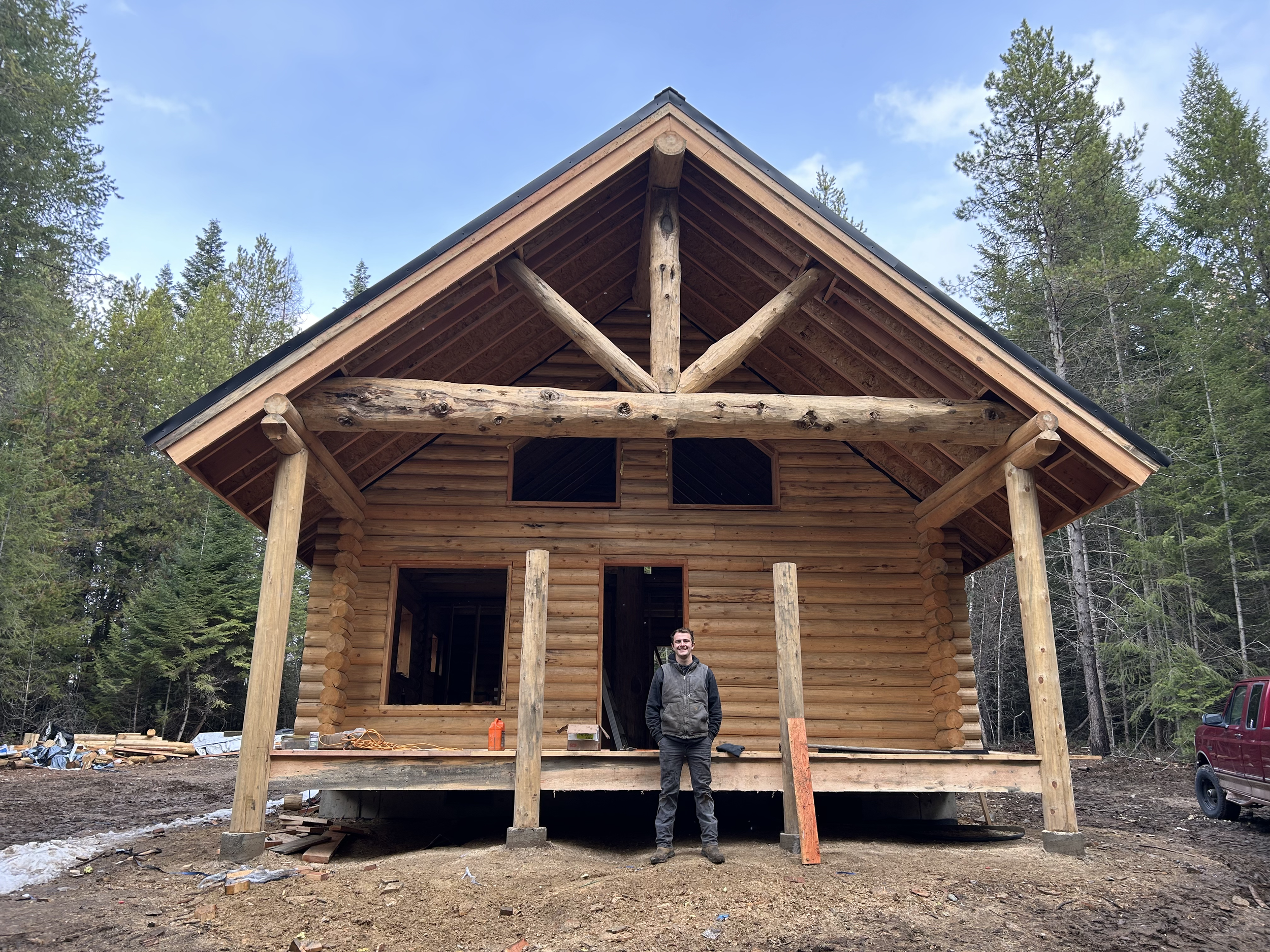 A man stands in front of a partially constructed wooden house in a forested area with trees on either side and blue sky above. The house has a peaked roof and logs for walls. There are construction materials and tools on the ground.