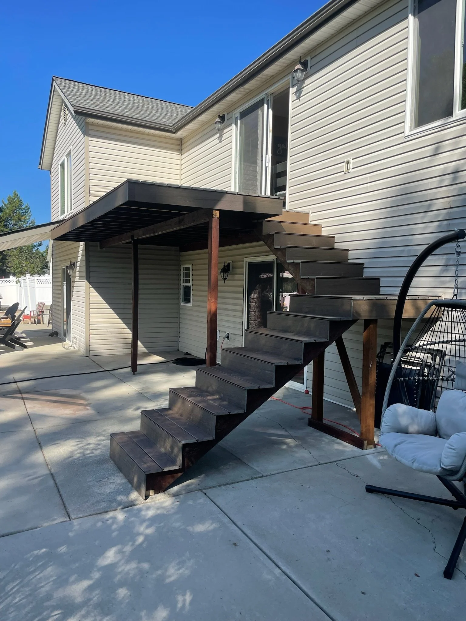 Backyard showing a two-story house with white siding, outdoor stairs with dark wood steps and a small balcony, concrete patio, and outdoor furniture under sunny sky.