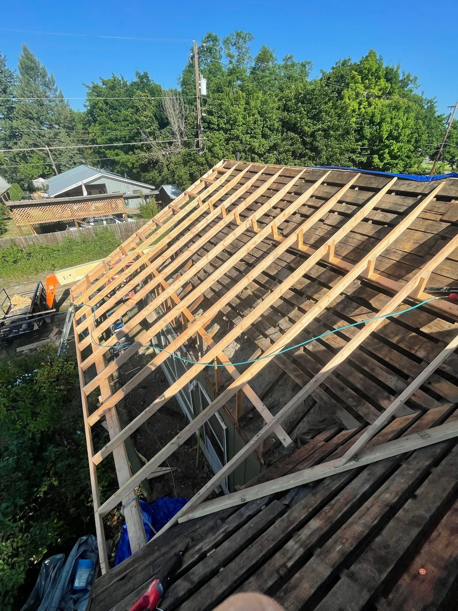 Construction site showing the framework of a roof under construction with wooden beams, partially covered with weathered shingles, and a clear blue sky overhead.