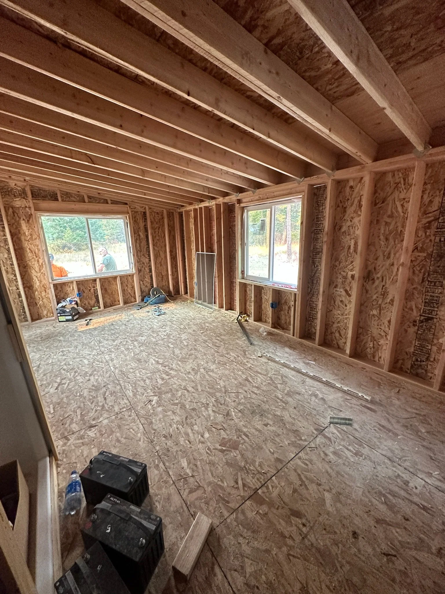 Interior of a house under construction with exposed wooden framing, OSB sheet walls, and two windows. Construction tools and materials are scattered on the unfinished plywood subfloor.