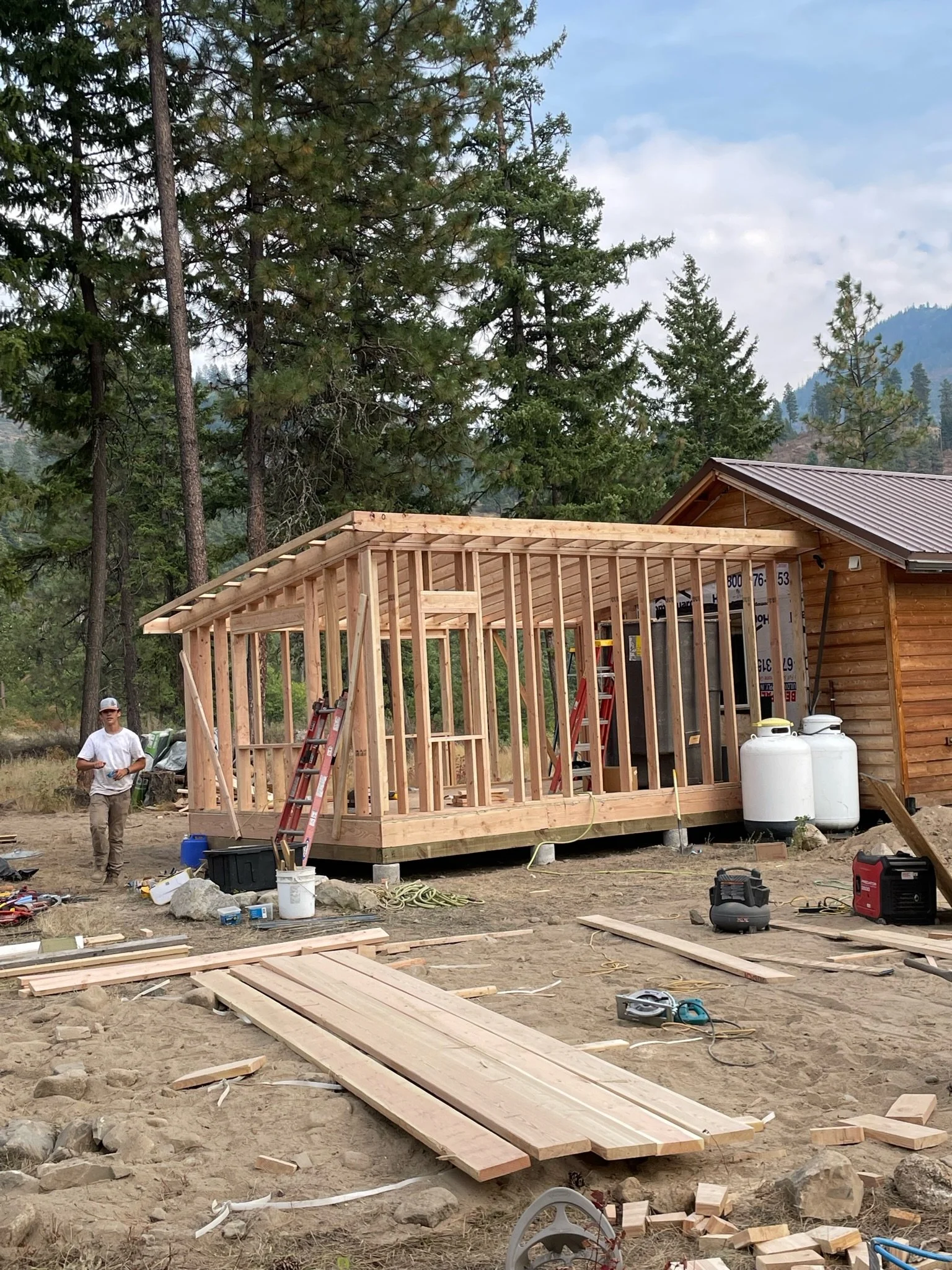 Construction site with a wooden building frame and a man standing near tools and materials, surrounded by trees and mountains in the background.