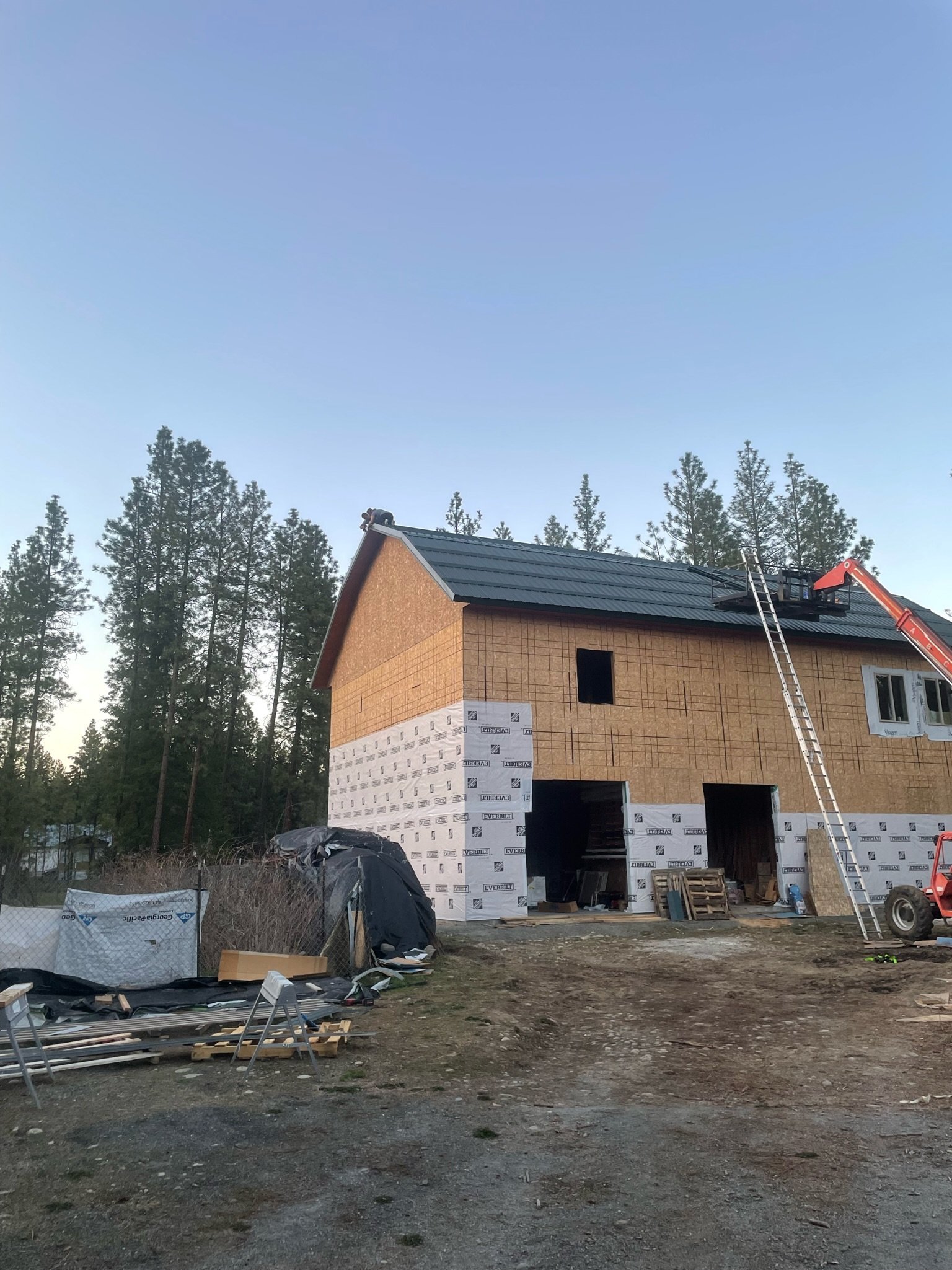 A house under construction with a ladder leaning against the roof, surrounded by construction materials and debris, with a forest of pine trees in the background.