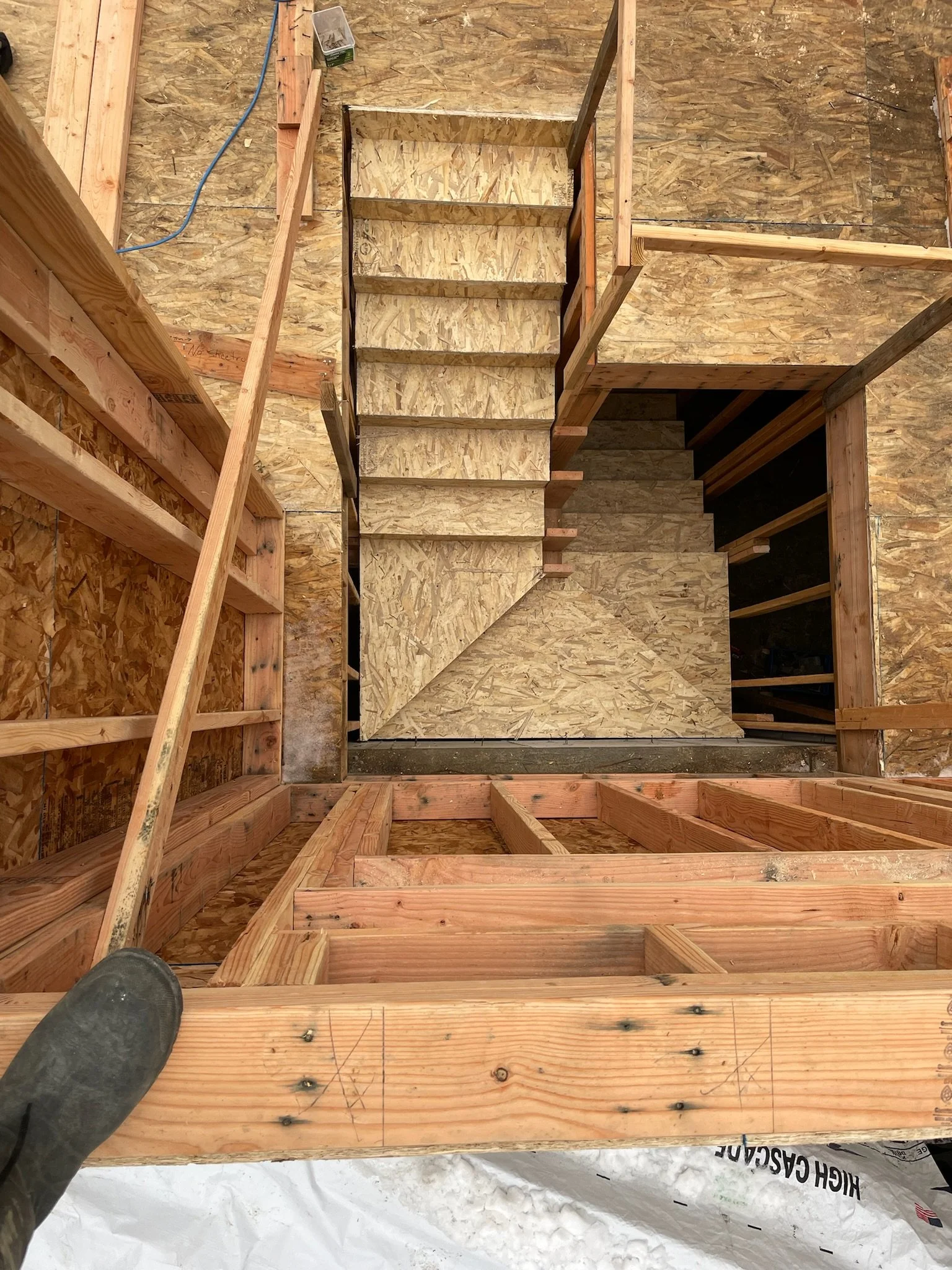 View from the top of a stairwell under construction, showing wooden framing and OSB plywood stairs and walls.