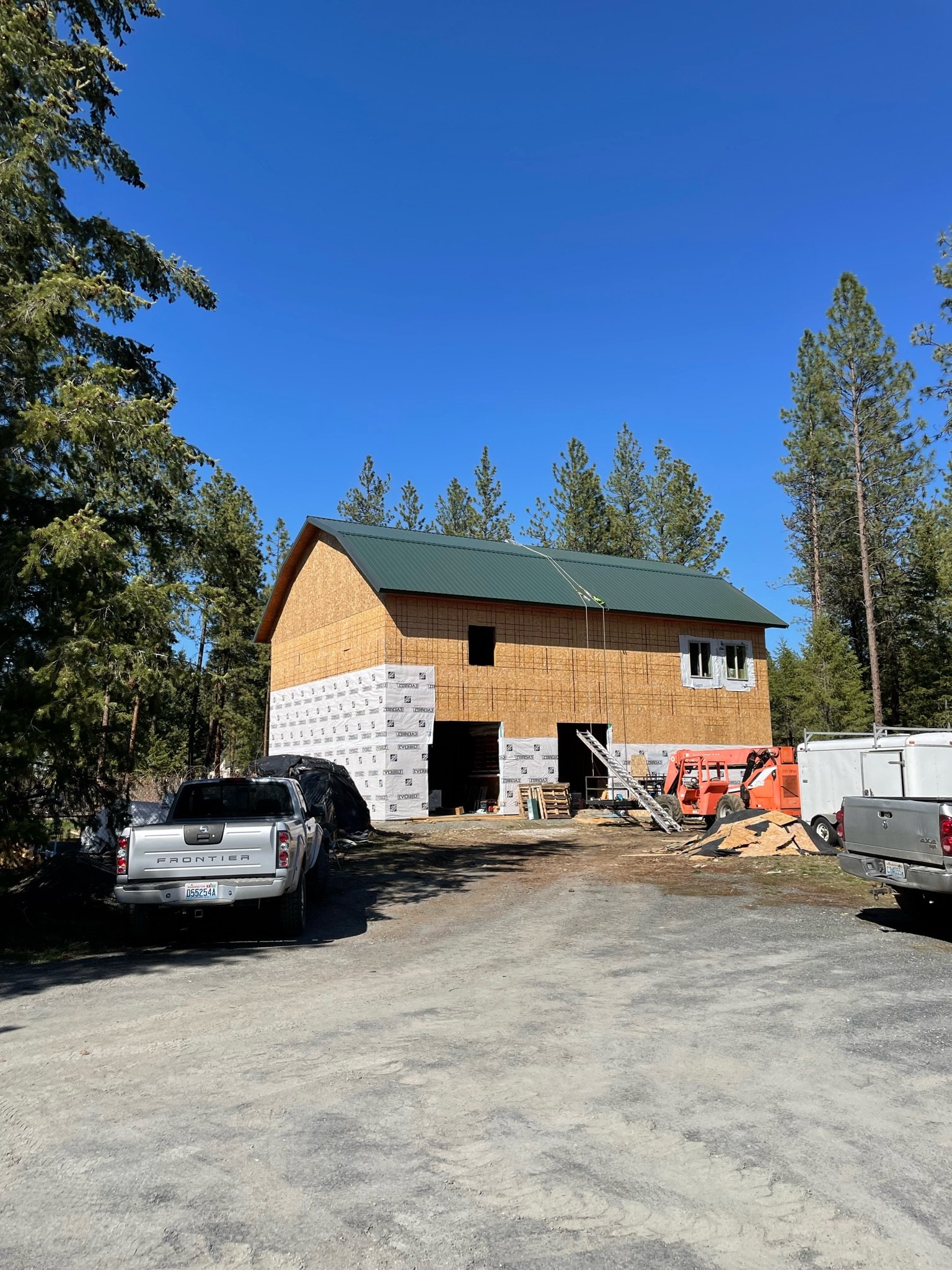 Construction site with a partially built house, construction vehicles, and parked cars, surrounded by tall trees under a clear blue sky.