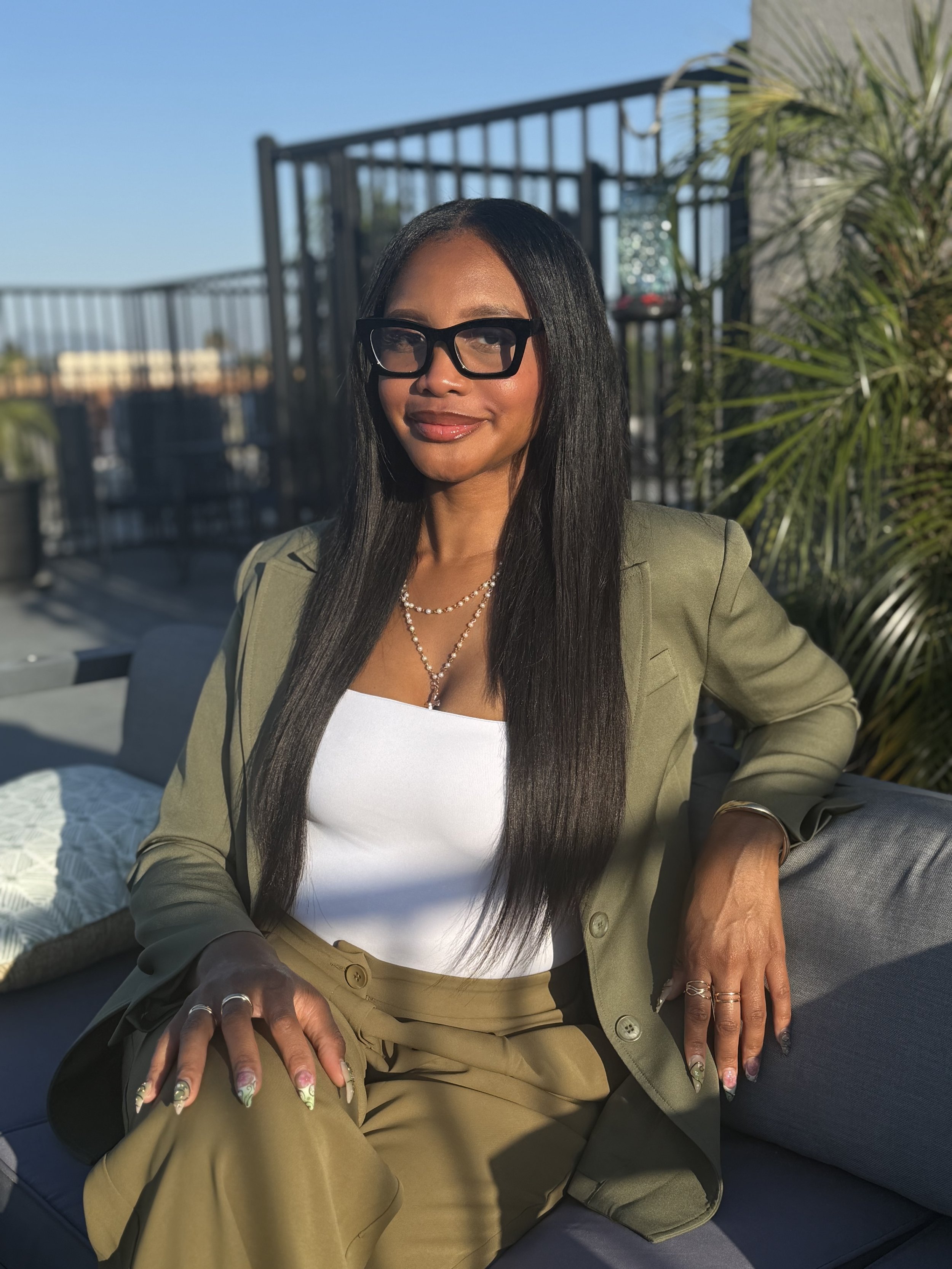 A woman with long black hair and glasses smiling while sitting outdoors on a patio with a black railing, green plants, and a clear blue sky in the background.