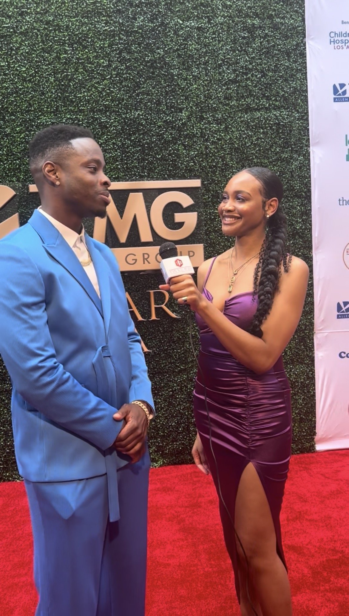 A woman holding a microphone interviewing a man in a blue suit on a red carpet at a formal event.