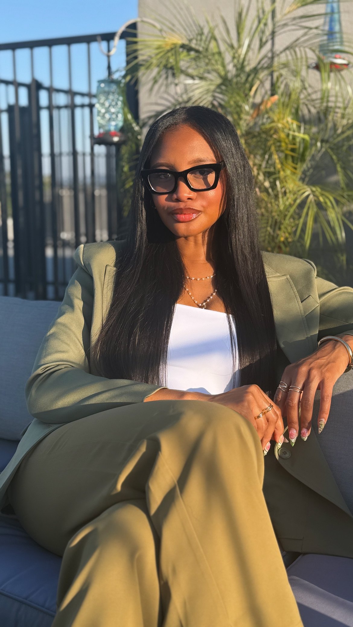 A woman with long black hair, wearing black glasses, a green blazer, and beige pants, sitting outdoors near a plant with sunlight on her face.