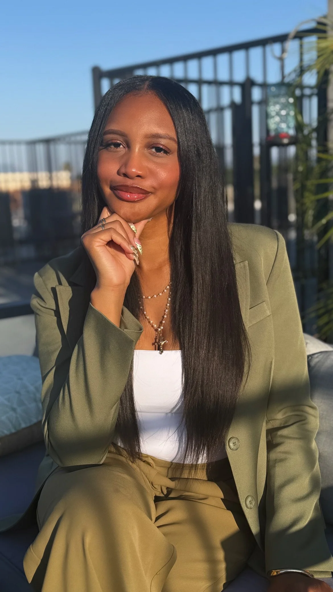 A woman with long dark hair and a confident expression, sitting outdoors on a sunny day. She is wearing an olive green blazer and beige pants, with layered necklaces. The background shows a black metal fence and a clear blue sky.