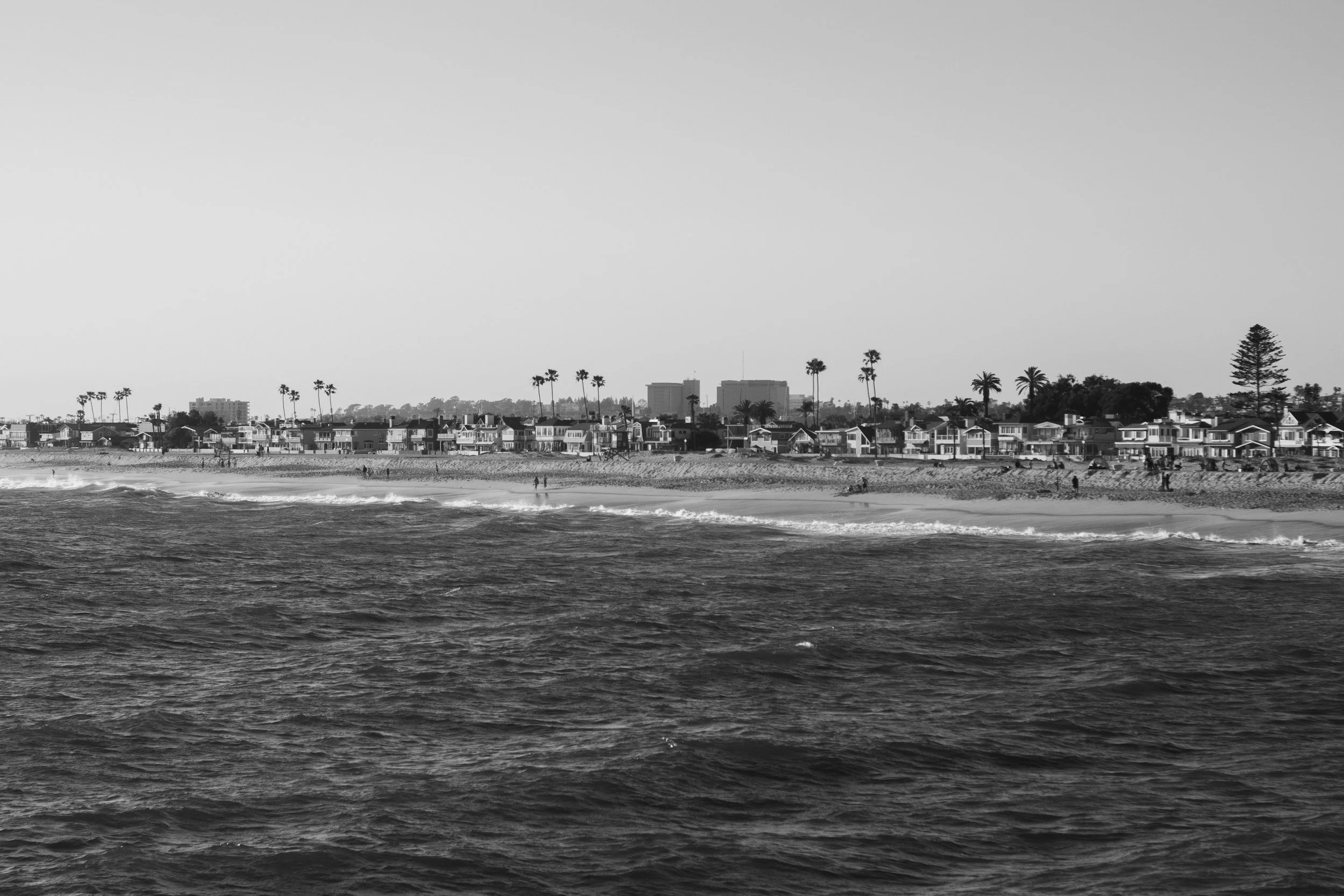 Black and white photo of a coastal beach with waves in the foreground, houses along the shoreline, and palm trees and buildings in the background.