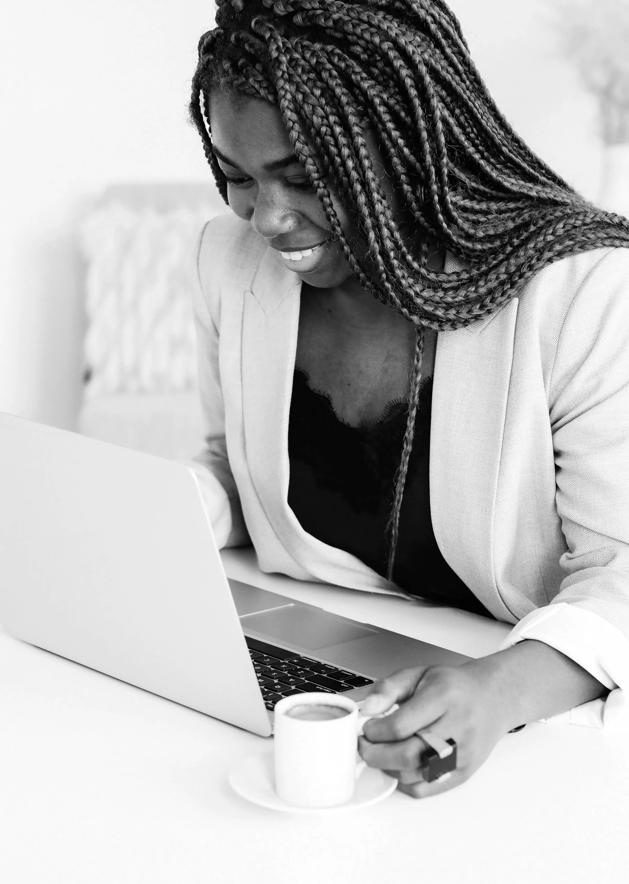 A smiling woman with braided hair using a laptop at a desk, holding a small cup of coffee or espresso.