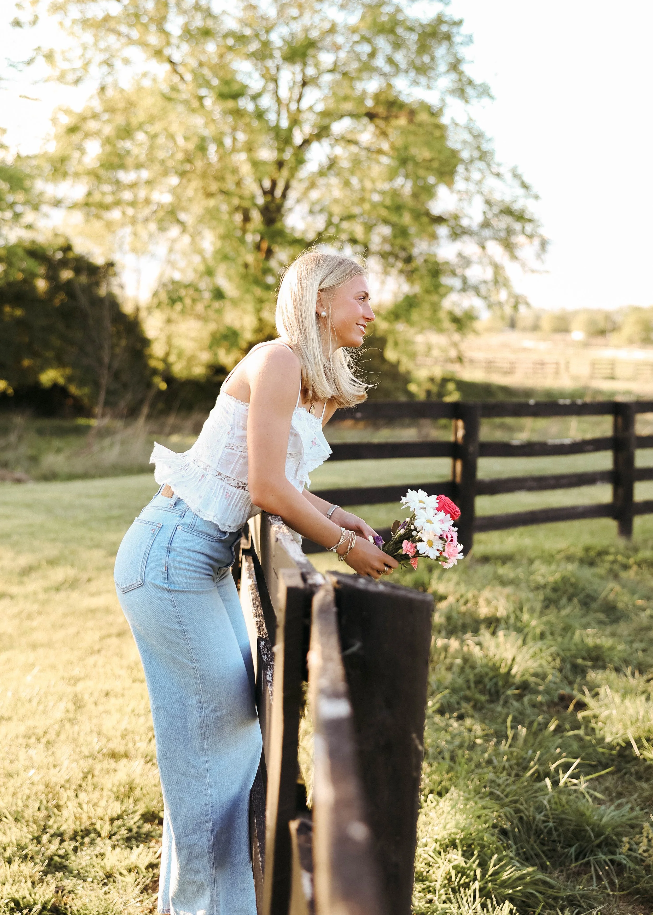 Senior girl posing by a wooden fence at the Kentucky Horse Park in Lexington, KY, surrounded by beautiful trees and open green fields.