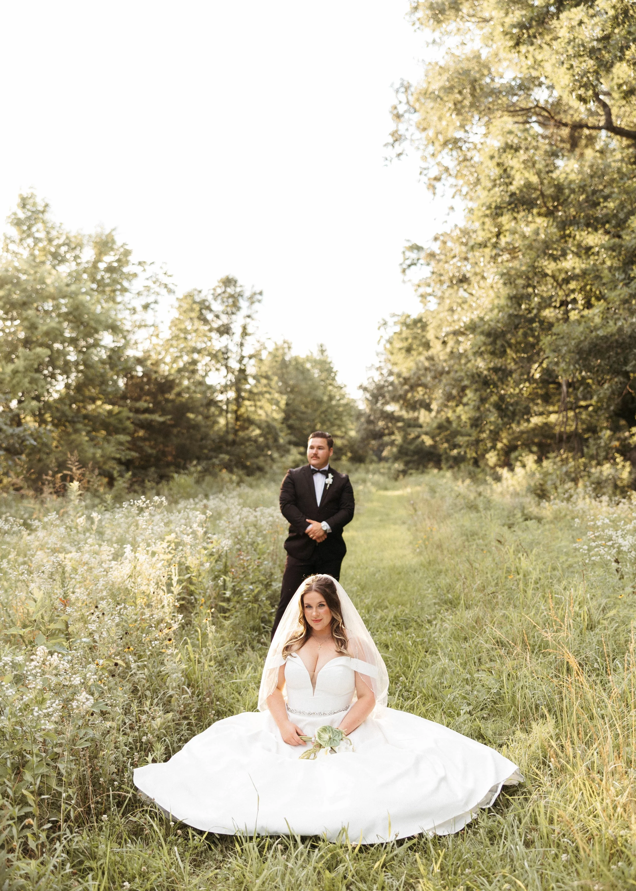A bride is sitting in a wildflower field in Kentucky while her groom is behind her staring into the fields