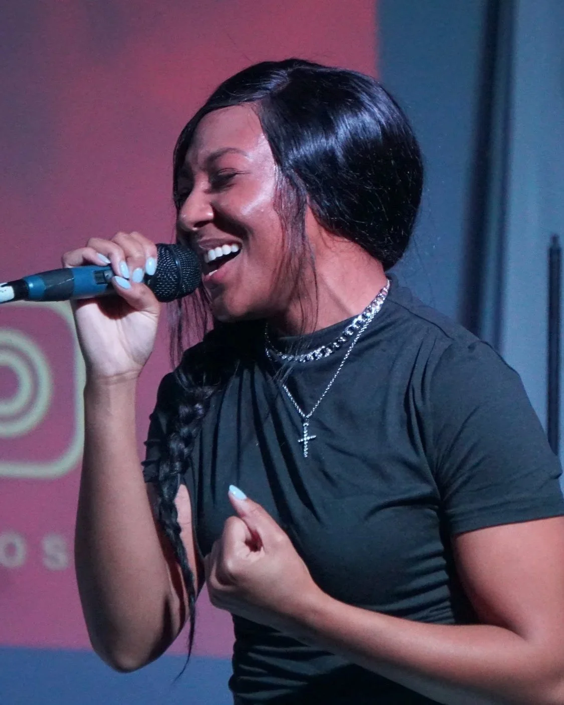 A woman singing passionately into a microphone, with her eyes closed and her hand clenched near her chest, wearing a black shirt and layered necklaces.