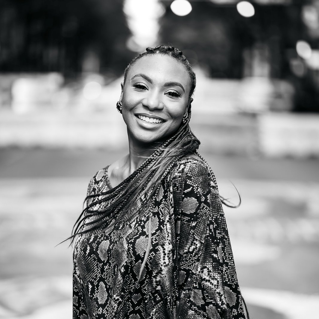 Black and white photo of a smiling woman with long braided hair, wearing earrings and a patterned top, outdoors in a blurred background