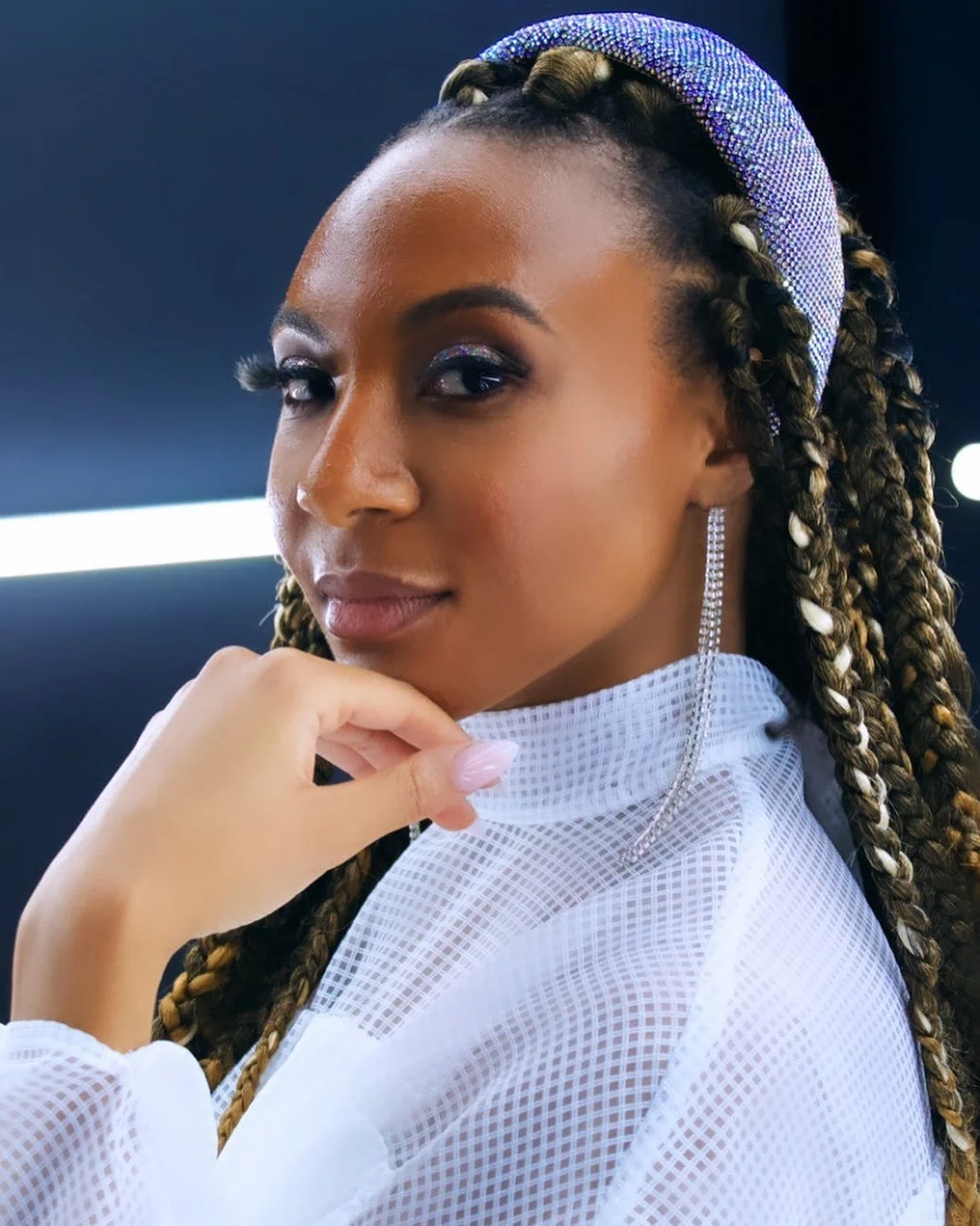 Close-up of a woman with braids wearing a sparkling headband, long dangling earrings, and a white mesh top, touching her chin with her hand.