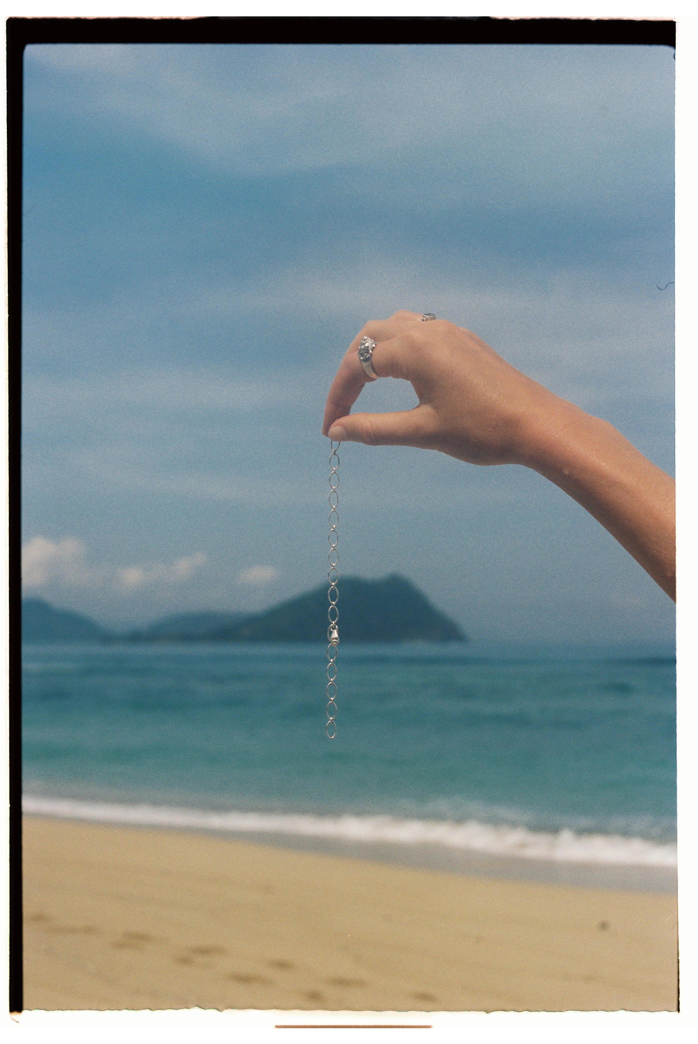 A woman's hand holding a silver chain with an ocean and island in the background.
