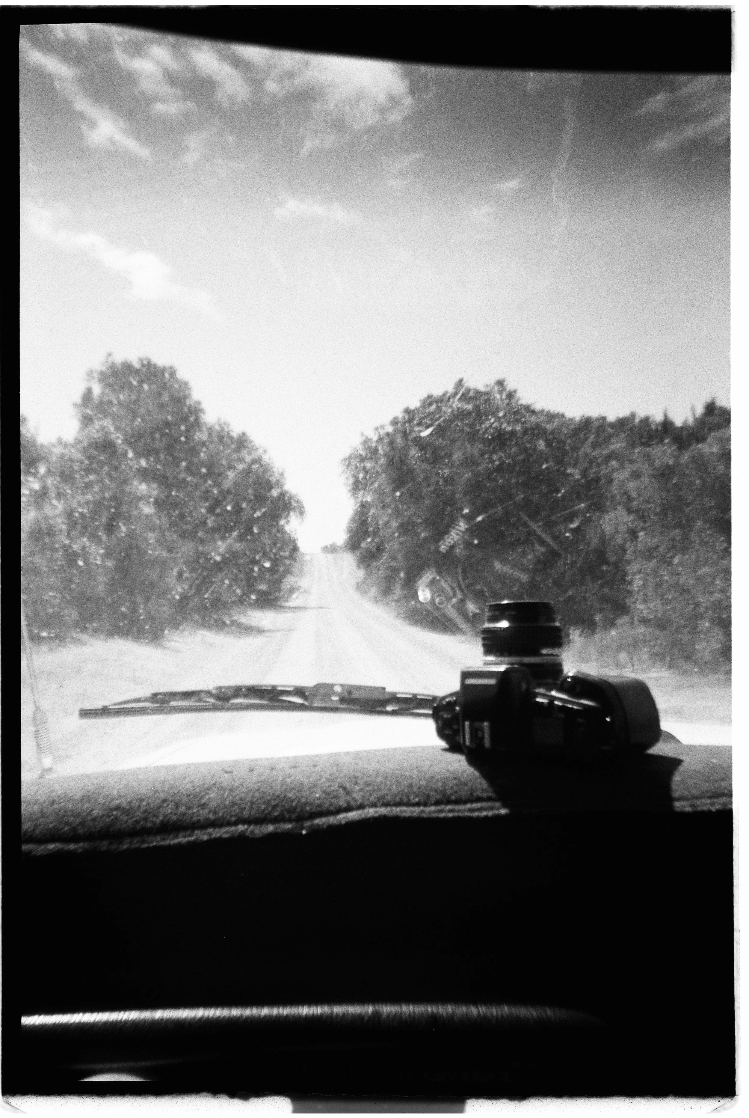 View from inside a vehicle looking through the windshield at a dirt road flanked by trees on both sides, with clouds in the sky. There is a camera placed on the dashboard.