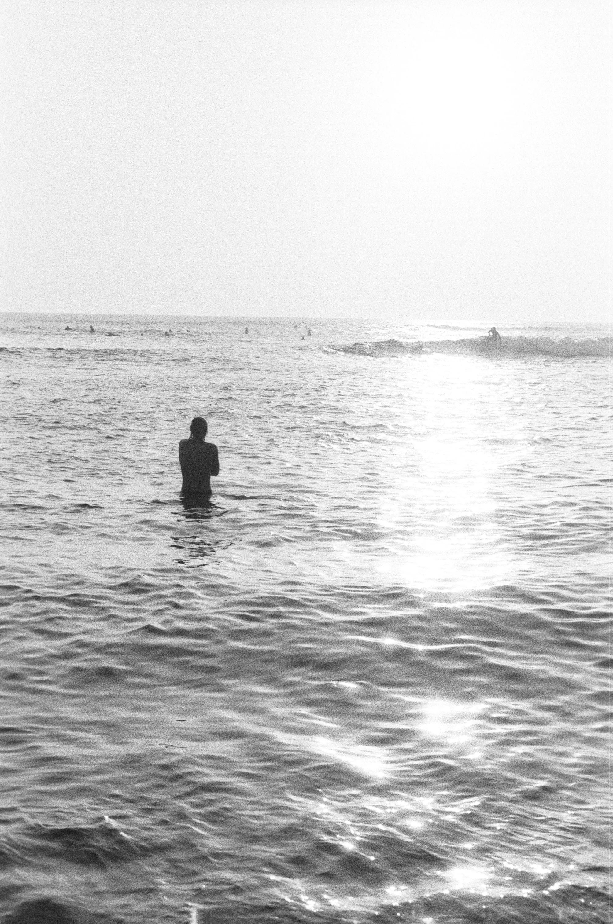 Silhouette of a person standing in the ocean with other surfers in the background, bright sunlight reflecting on the water.