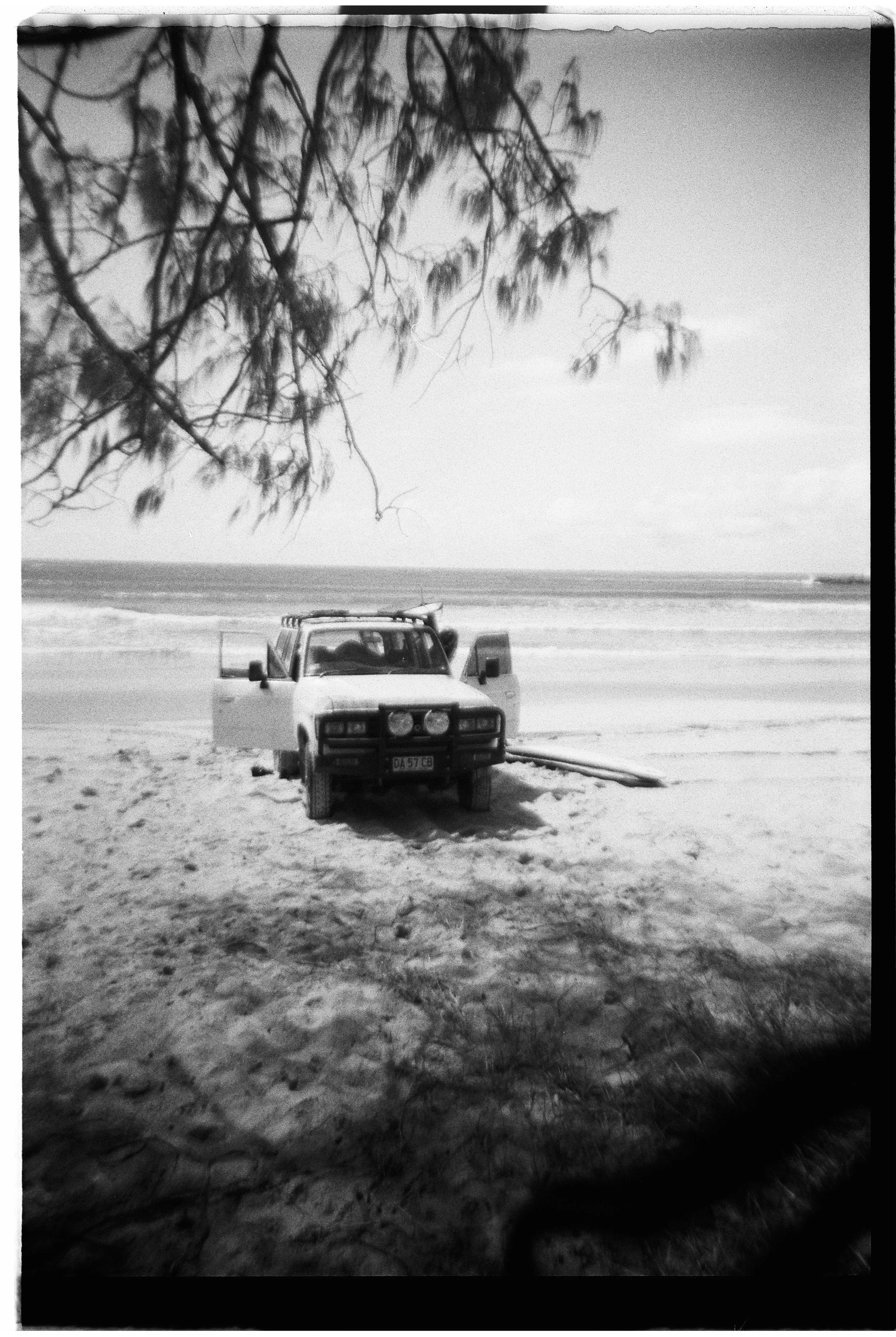 A black and white photo of a vehicle parked on a sandy beach near the ocean with an open door, under a tree with overhanging branches.