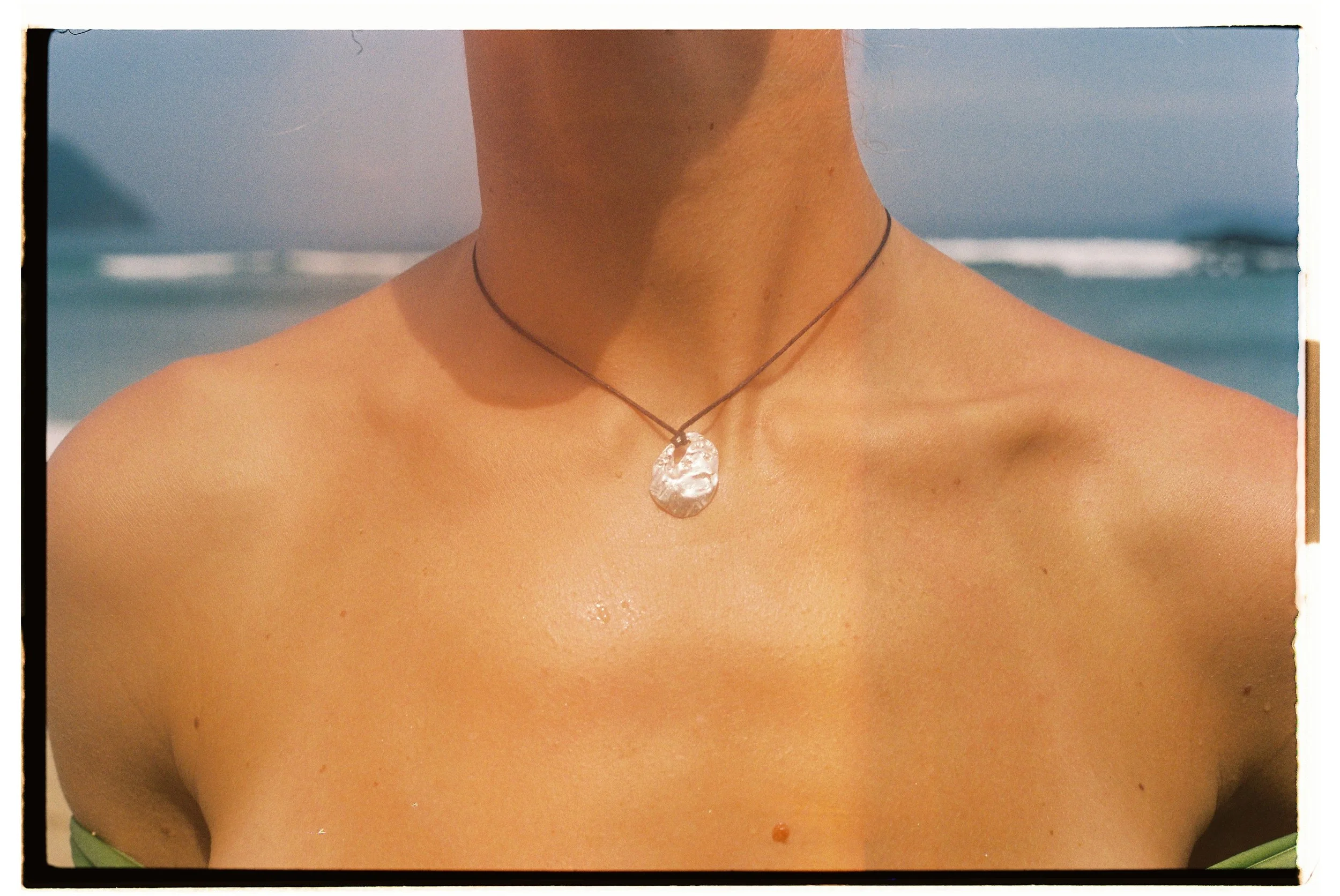 Close-up of a woman's bare shoulders and neck wearing a necklace with a shell pendant at the beach with waves in the background.