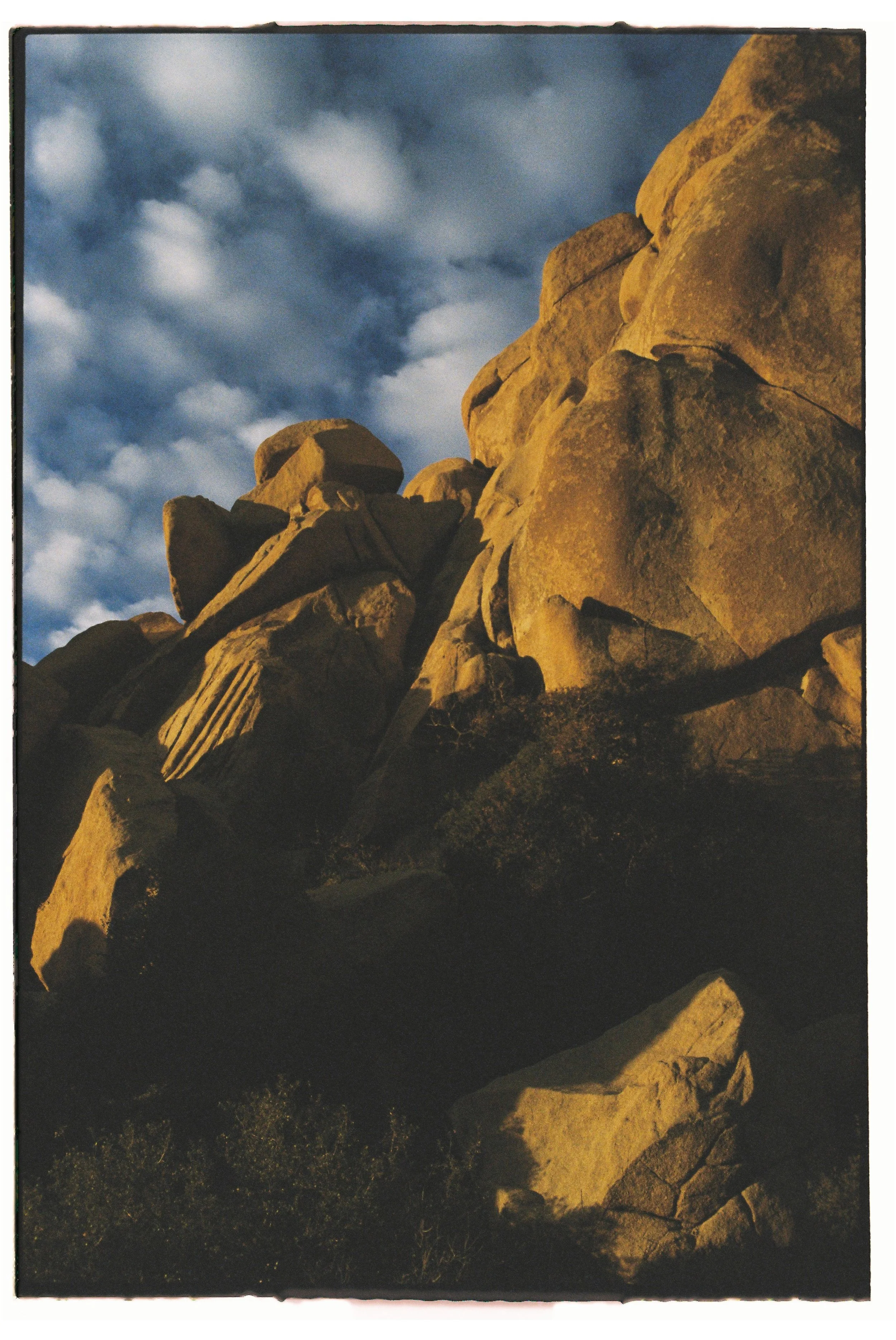 Sunlit large boulders and rocks on a mountainside against a cloudy sky at sunset.