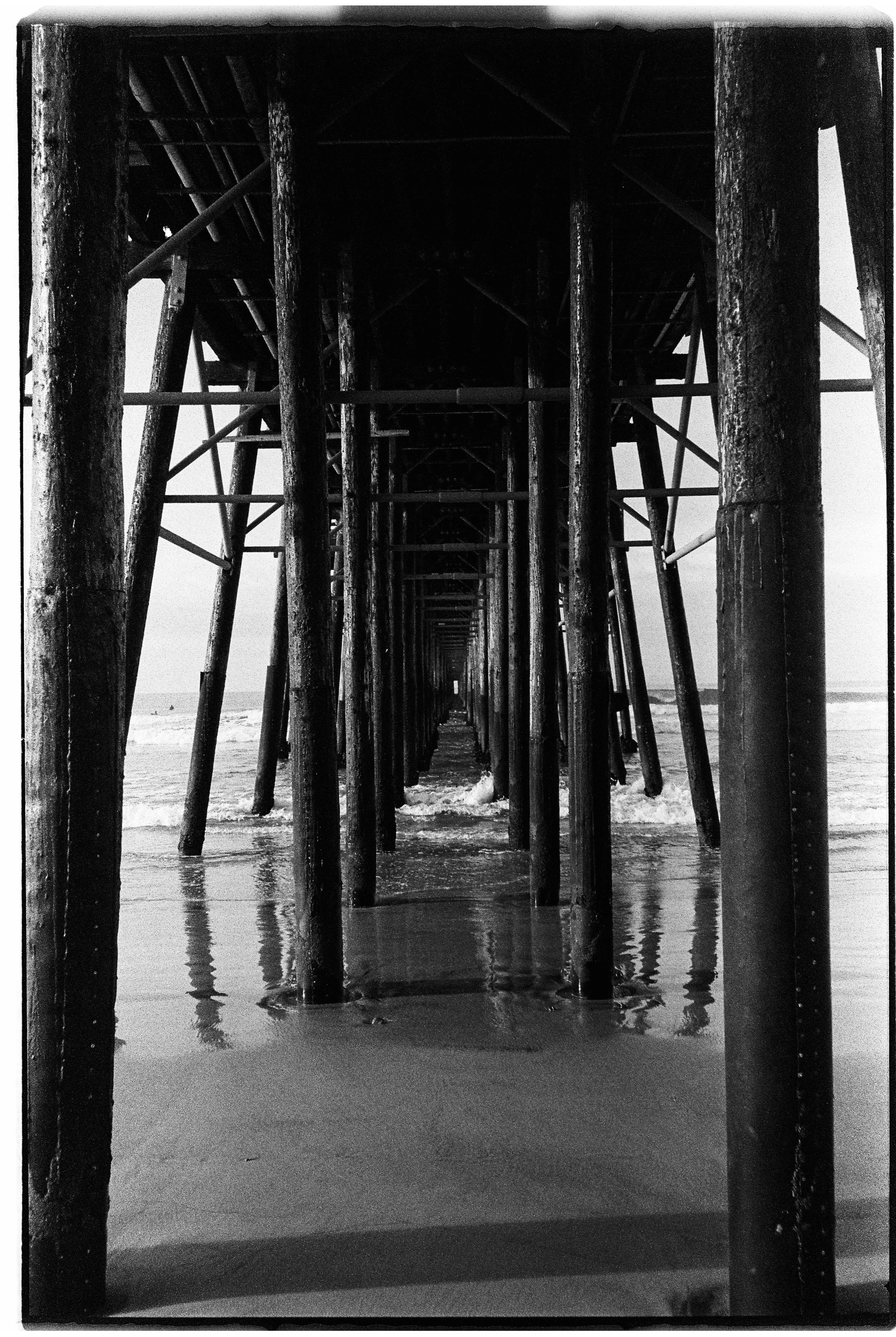 View of a pier extending over the ocean, taken from beneath, showing wooden support beams and the sandy beach below in black and white.