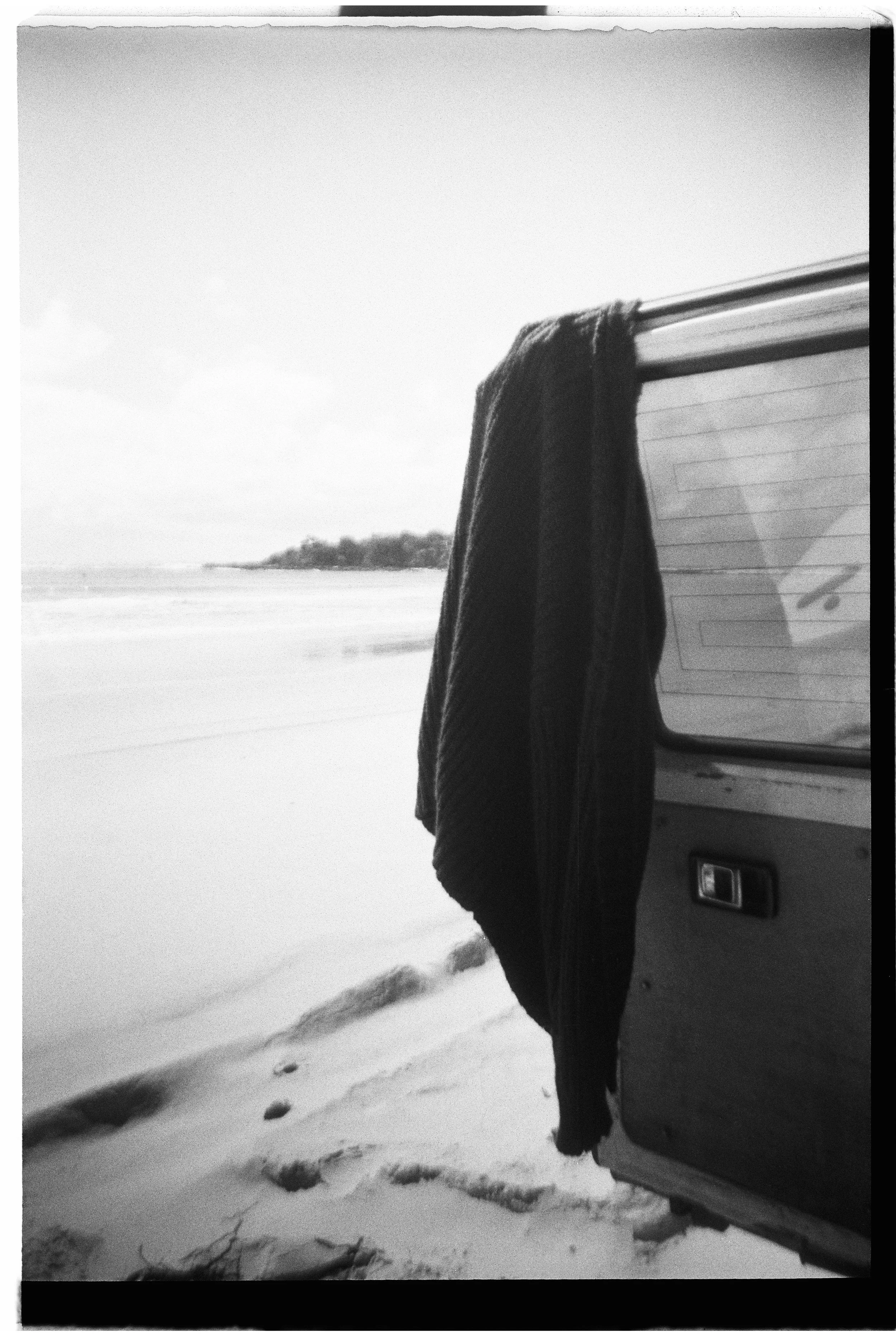 Clothes hanging on the side of a van parked on a beach with waves and a small landmass in the background, black and white photo.