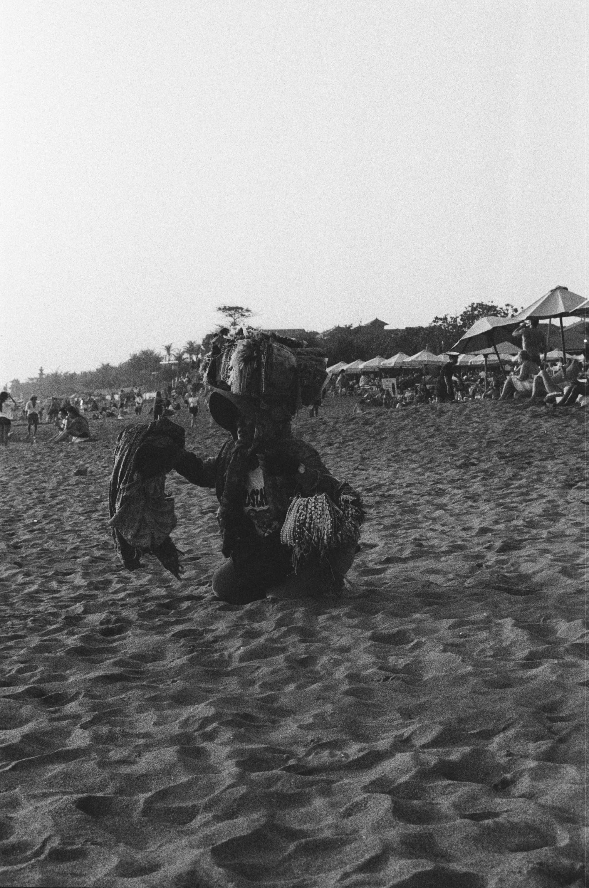 A person kneeling on a sandy beach, carrying a bundle on their head, with other beachgoers and umbrellas in the background.