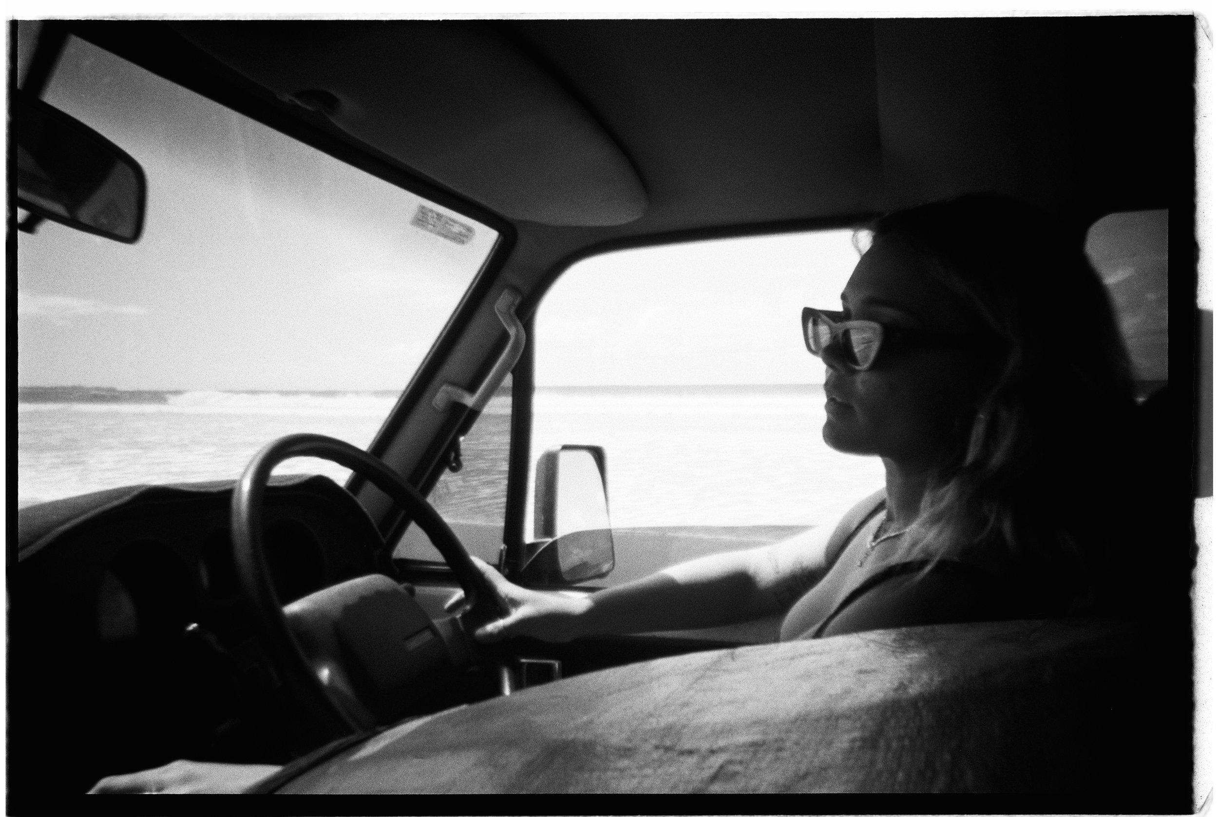 A woman driving a vehicle with a view of water through the front window in black and white.