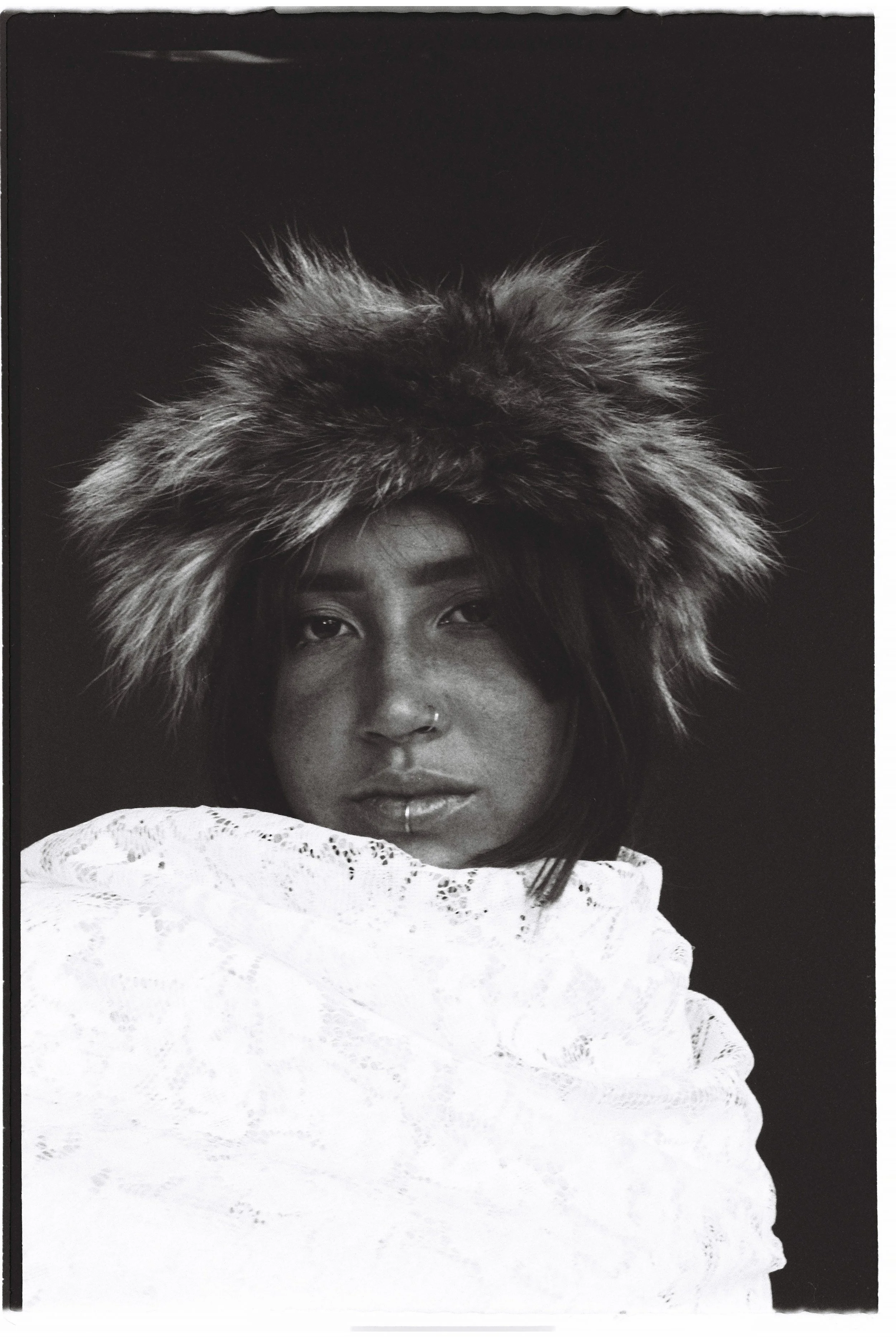 Black and white portrait of a woman wearing a large, furry hat and an intricate white lace garment, with dark hair and a piercing on her nose.