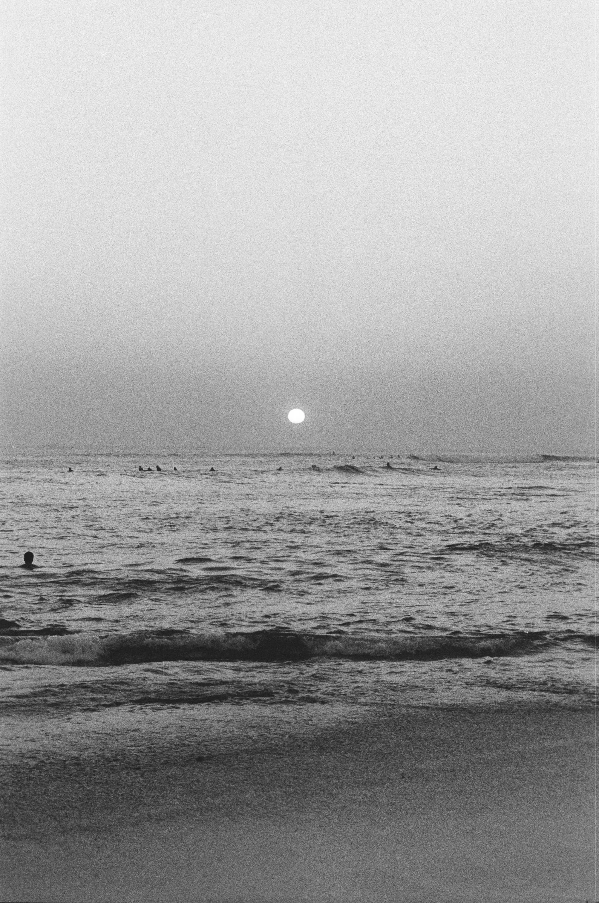 Black and white photo of a beach during sunset, with some people in the water and small waves on the shore.